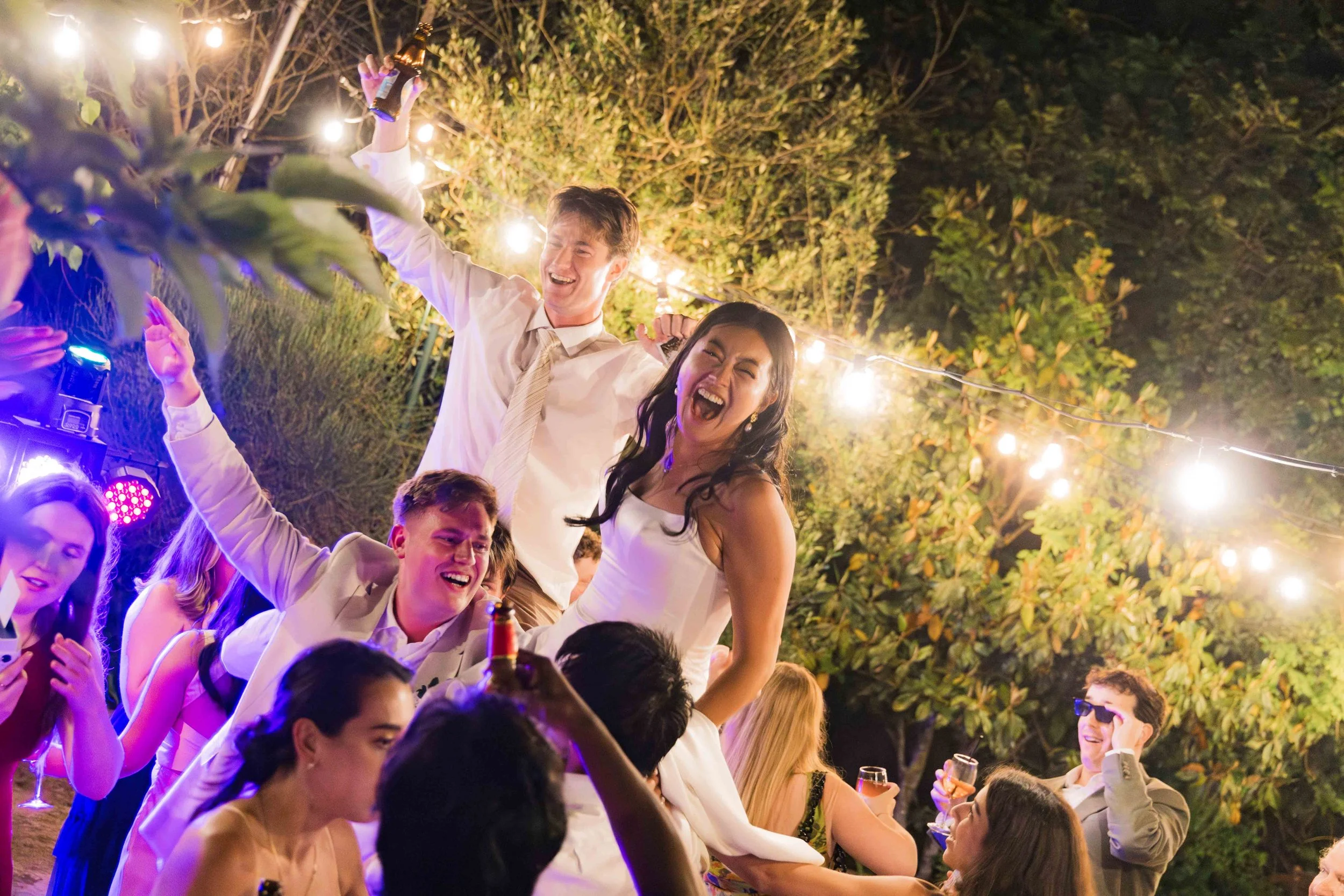 A group of people celebrating at an outdoor party at night, with string lights overhead. Two women and two men are standing, smiling and raising their drinks. Others sit at tables, socializing and laughing.