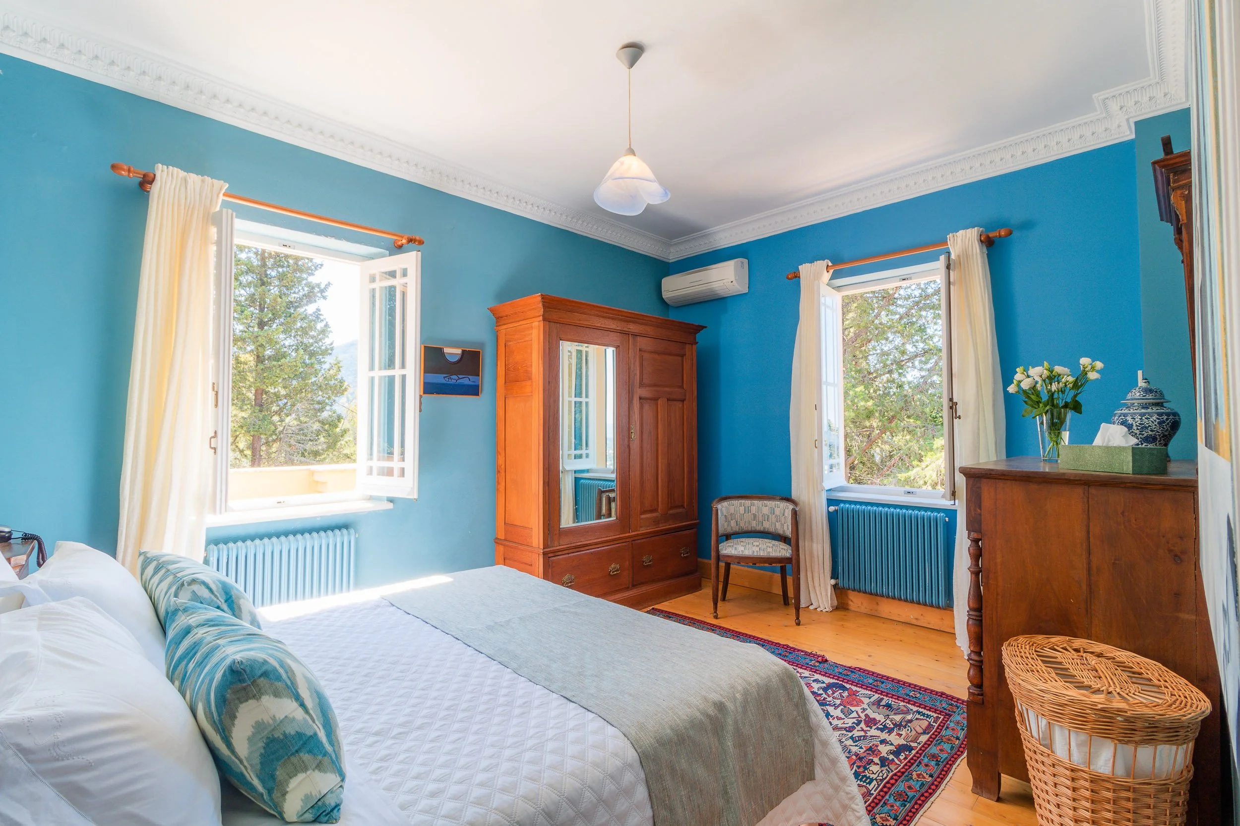 Bedroom with blue walls, white ceiling, and hardwood floor. There is a bed with white bedding and decorative pillows, a wooden wardrobe with a mirror, a wooden dresser with a vase and flowers, two open windows with white curtains, a small chair, an a