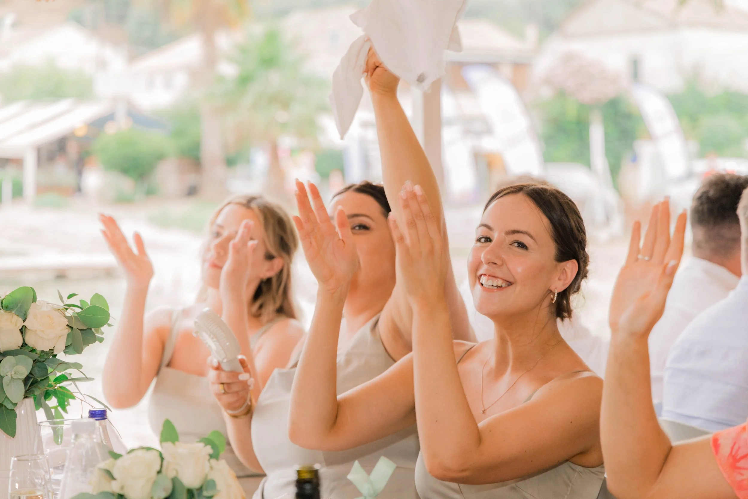 People celebrating at a wedding reception, smiling and throwing paper napkins in the air, with flowers and drinks on the table.