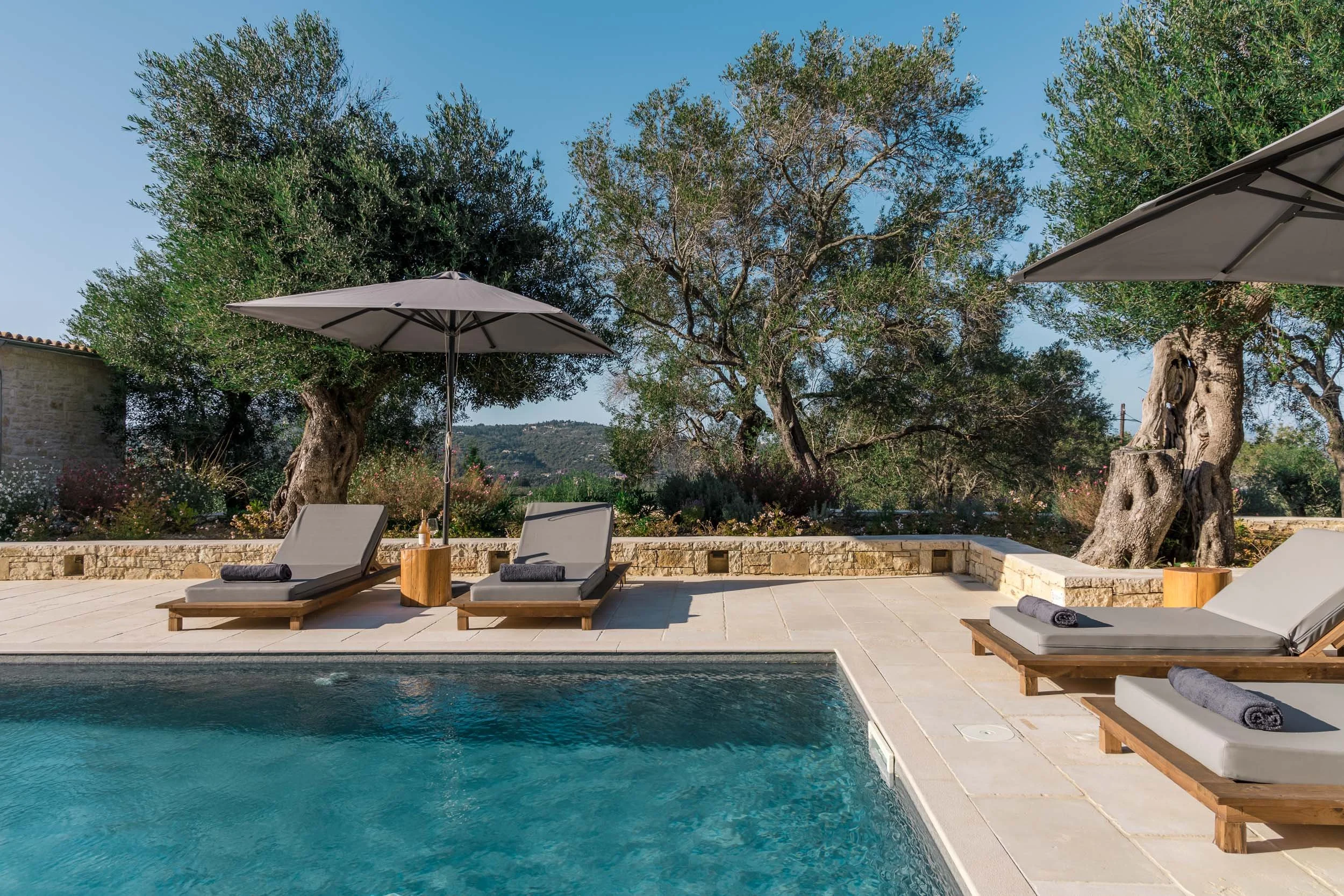 A swimming pool surrounded by a stone patio, with three lounge chairs and umbrella, trees in the background under a clear blue sky.