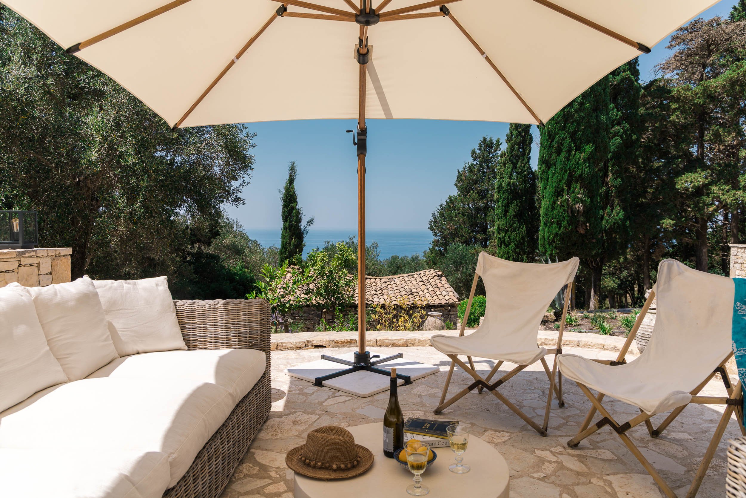 Outdoor patio area with white cushioned sofa, two white folding chairs, a large beige umbrella, and a small round table holding a straw hat, a bottle of wine, and two glasses of wine. The background features trees, a stone wall, a tiled roof, and a v