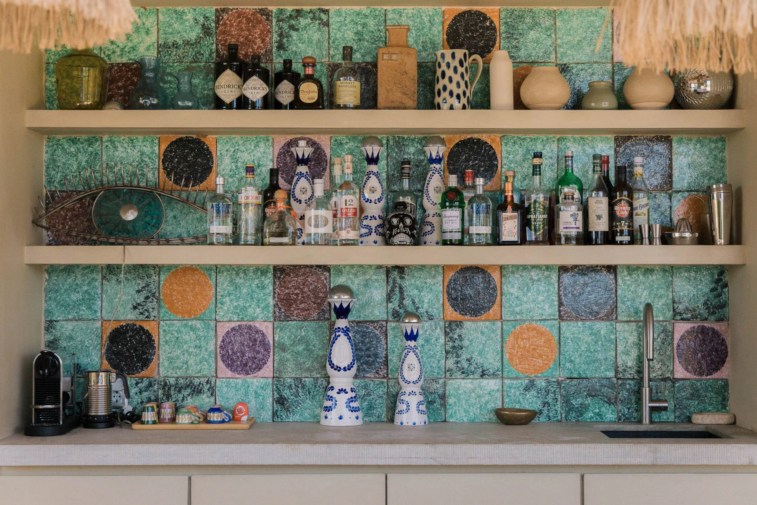 Colorful kitchen shelf with liquor bottles, colorful pottery, and decorative vases, set against a tiled wall with green, orange, black, and purple circles.