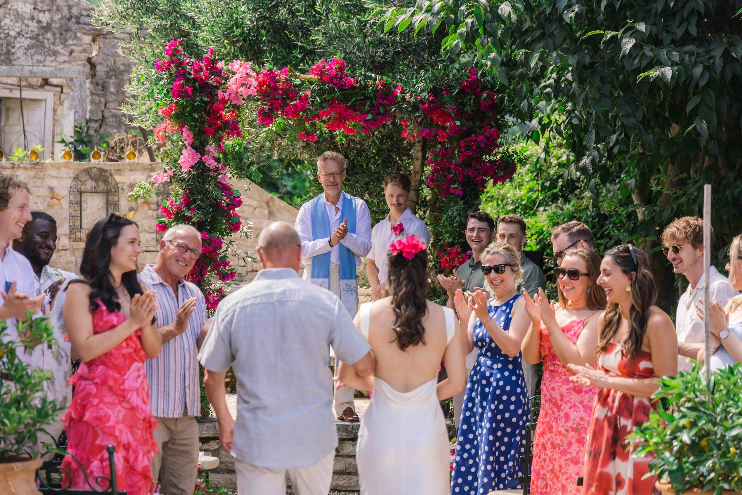 People at a wedding ceremony outdoors, with a bride and groom standing in front of an arch covered in pink and purple flowers, surrounded by friends and family clapping and smiling.