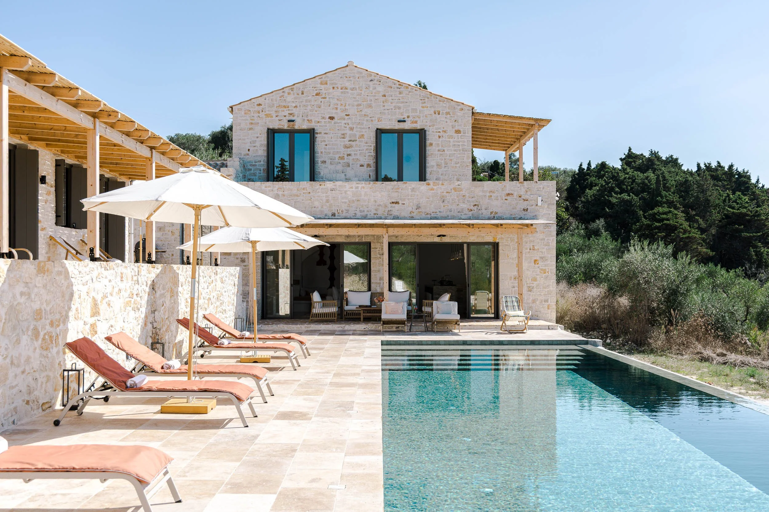 Luxury home with a swimming pool, surrounded by lounge chairs with pink cushions, white umbrellas, outdoor seating area, stone walls, and a backdrop of trees under a clear blue sky.