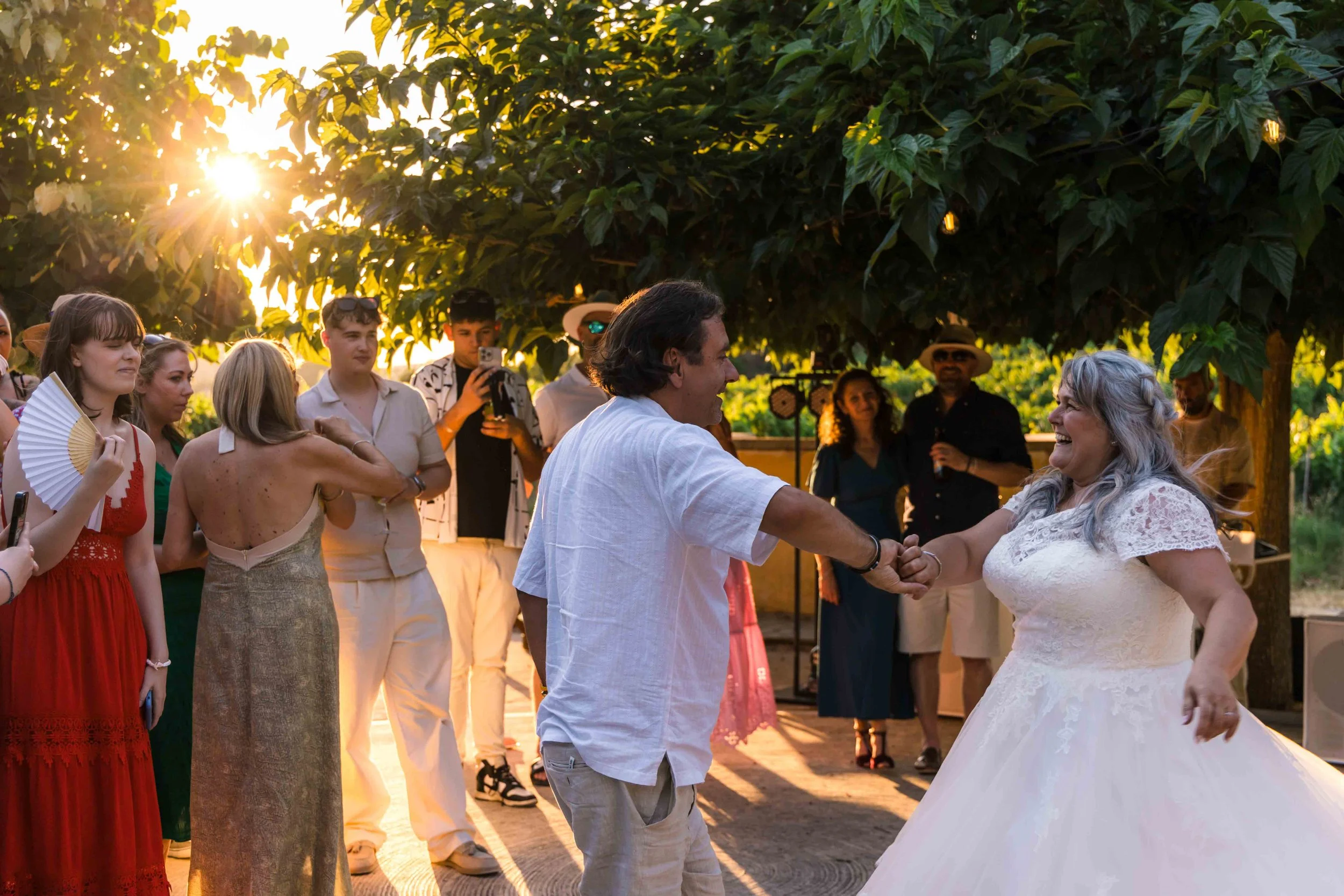 A wedding reception outdoors with a bride and groom dancing under a large tree during sunset, surrounded by friends and family.