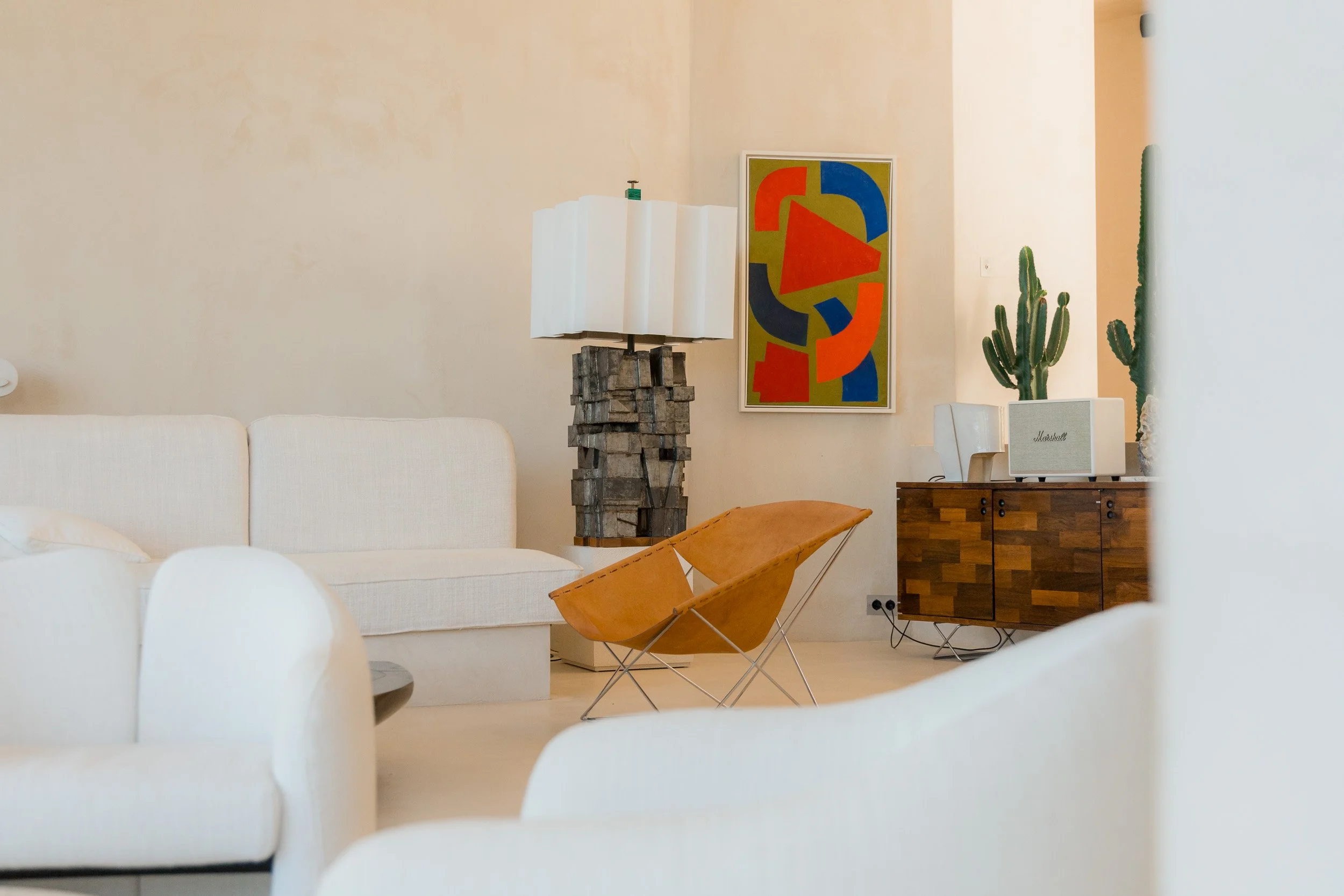 Modern living room with white sofa, orange lounge chair, wooden sideboard, cactus plants, colorful abstract artwork, and a unique lamp.