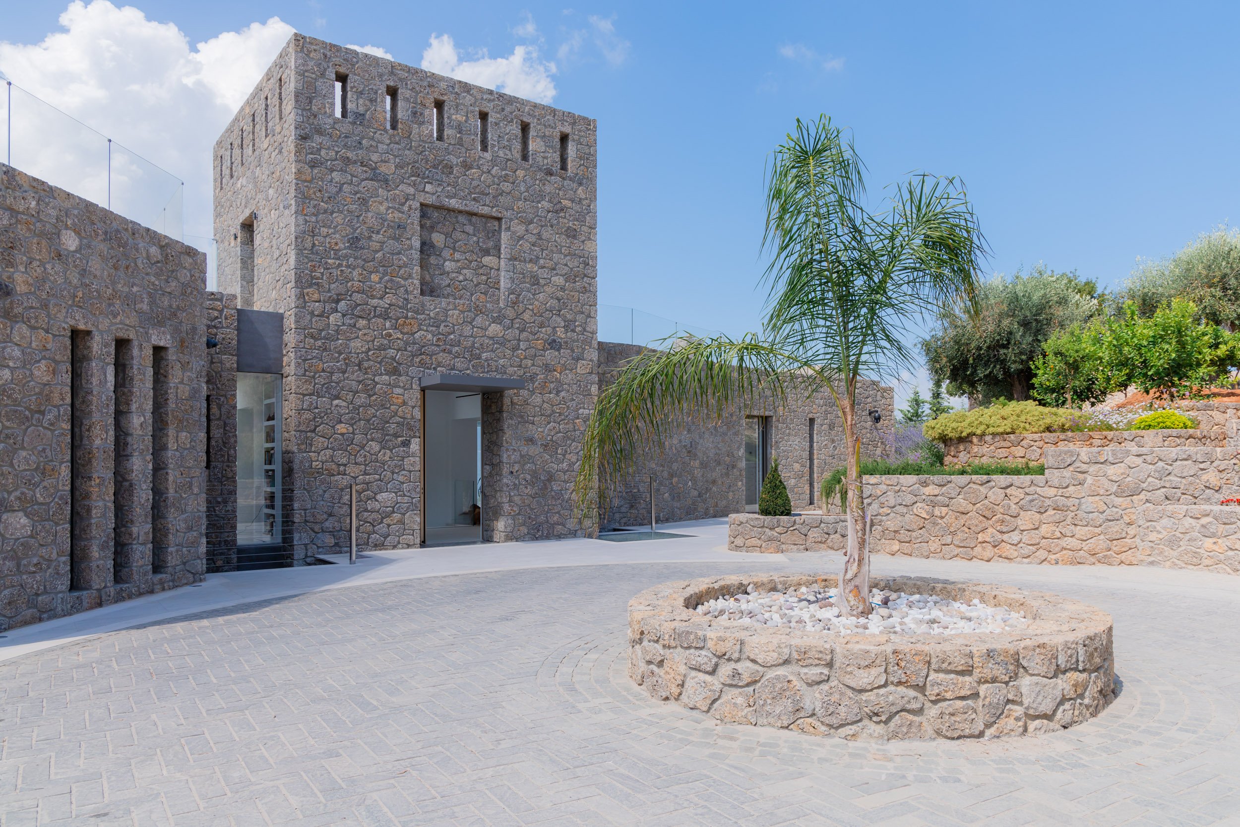 A modern stone building with a tower, surrounded by a landscaped courtyard with a small palm tree and various plants, under a blue sky with some white clouds.