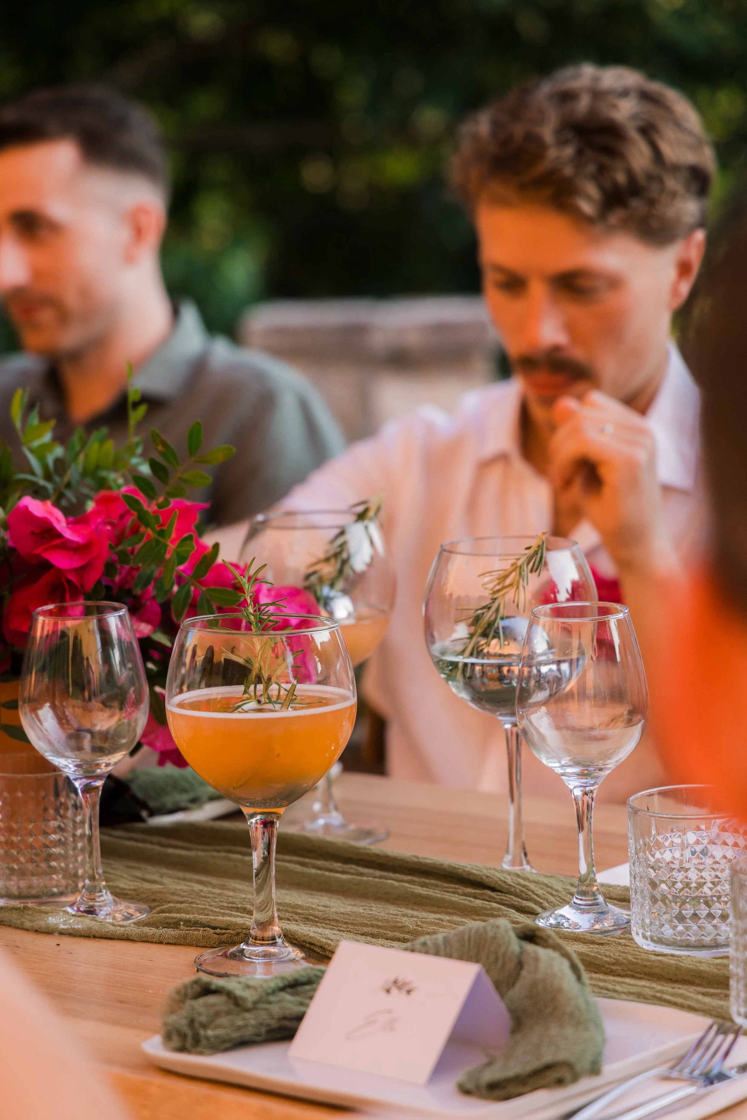 People sitting at a table outdoors with floral arrangements and wine glasses, enjoying a social event.