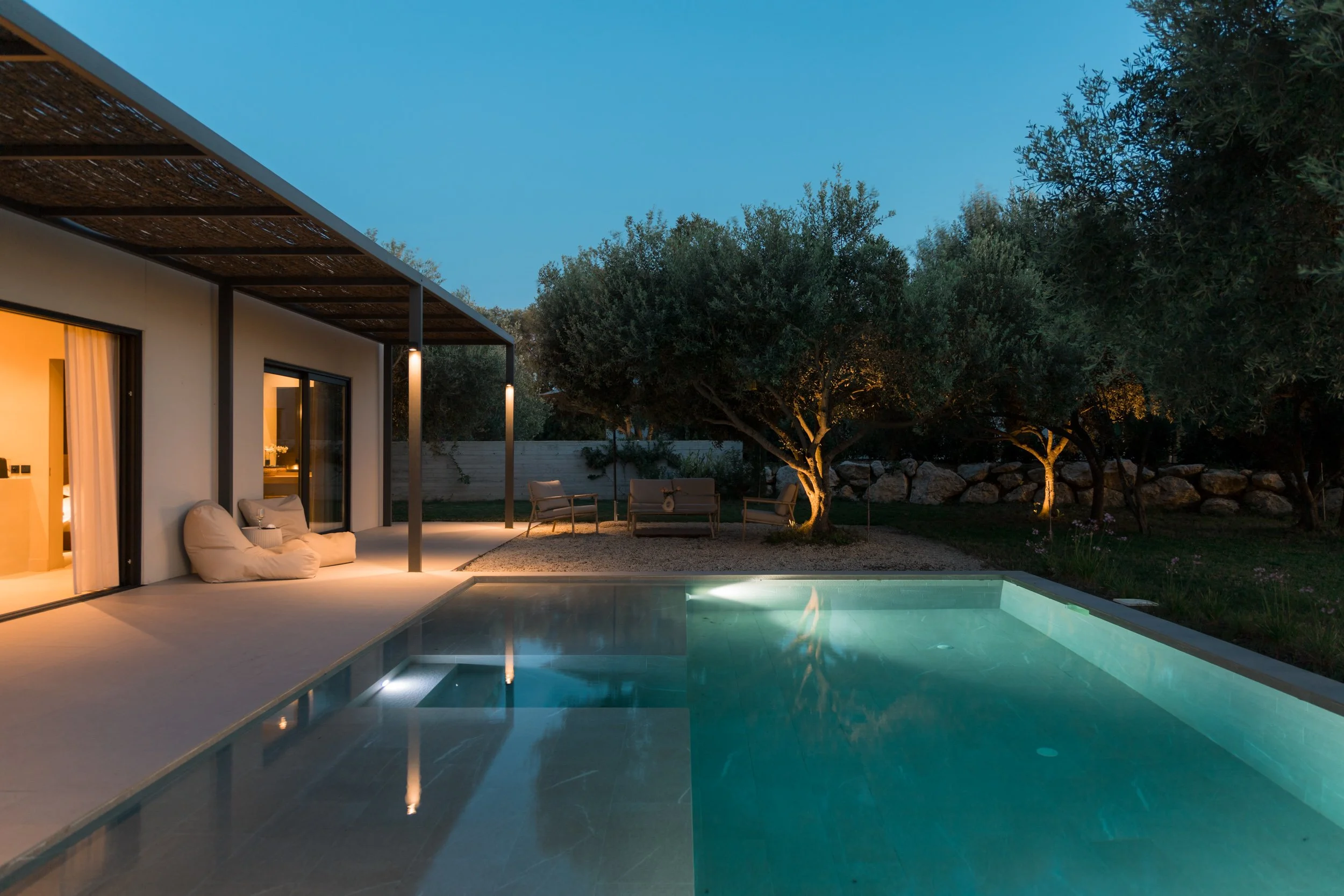 A modern backyard at dusk with a swimming pool, outdoor seating, and trees with lights illuminating the area.