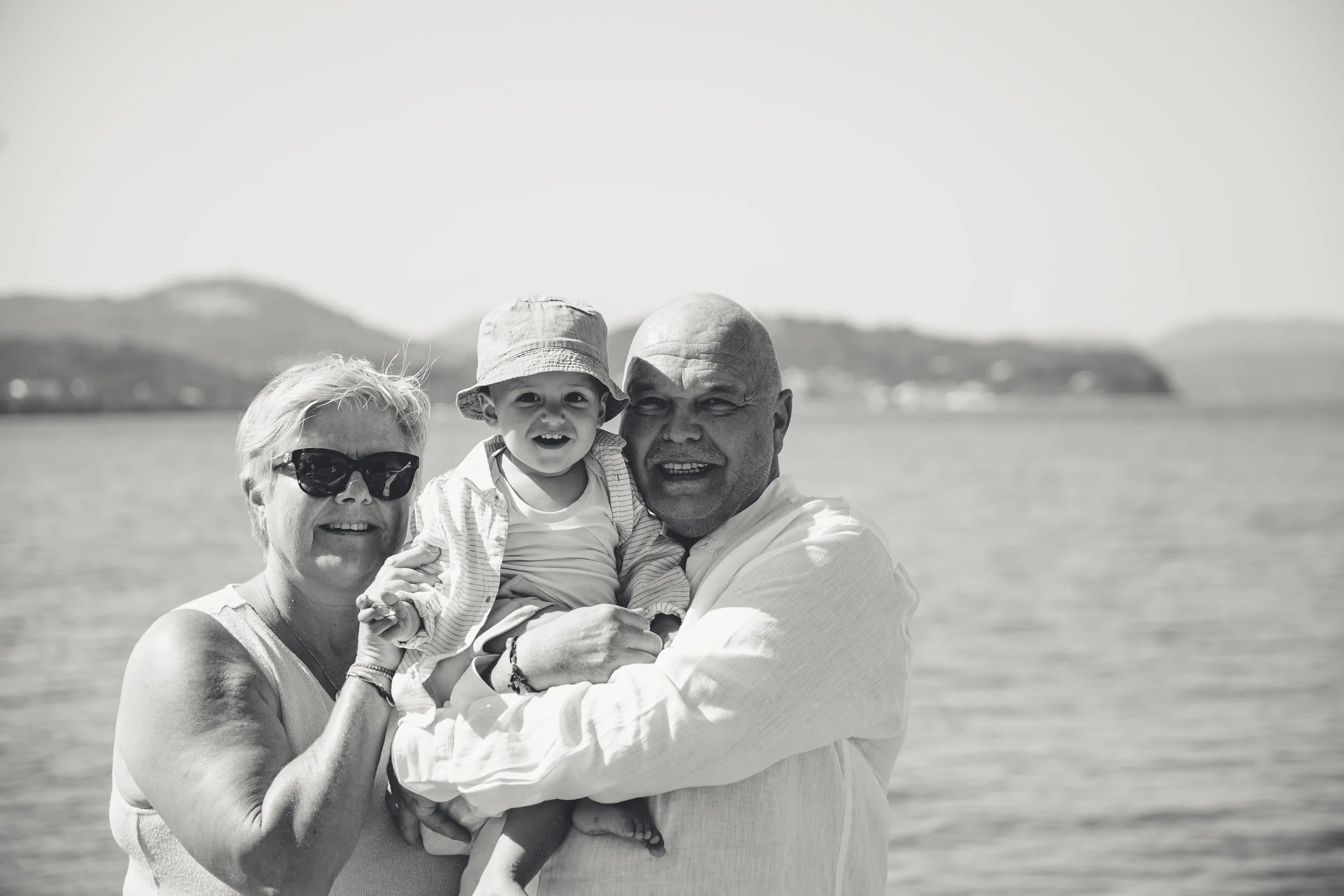 A family of three standing outdoors near a body of water, with mountains in the background. The woman wears sunglasses and a sleeveless top, holding a young child in a sunhat and striped shirt, while the man holds the child. They are smiling and enjo