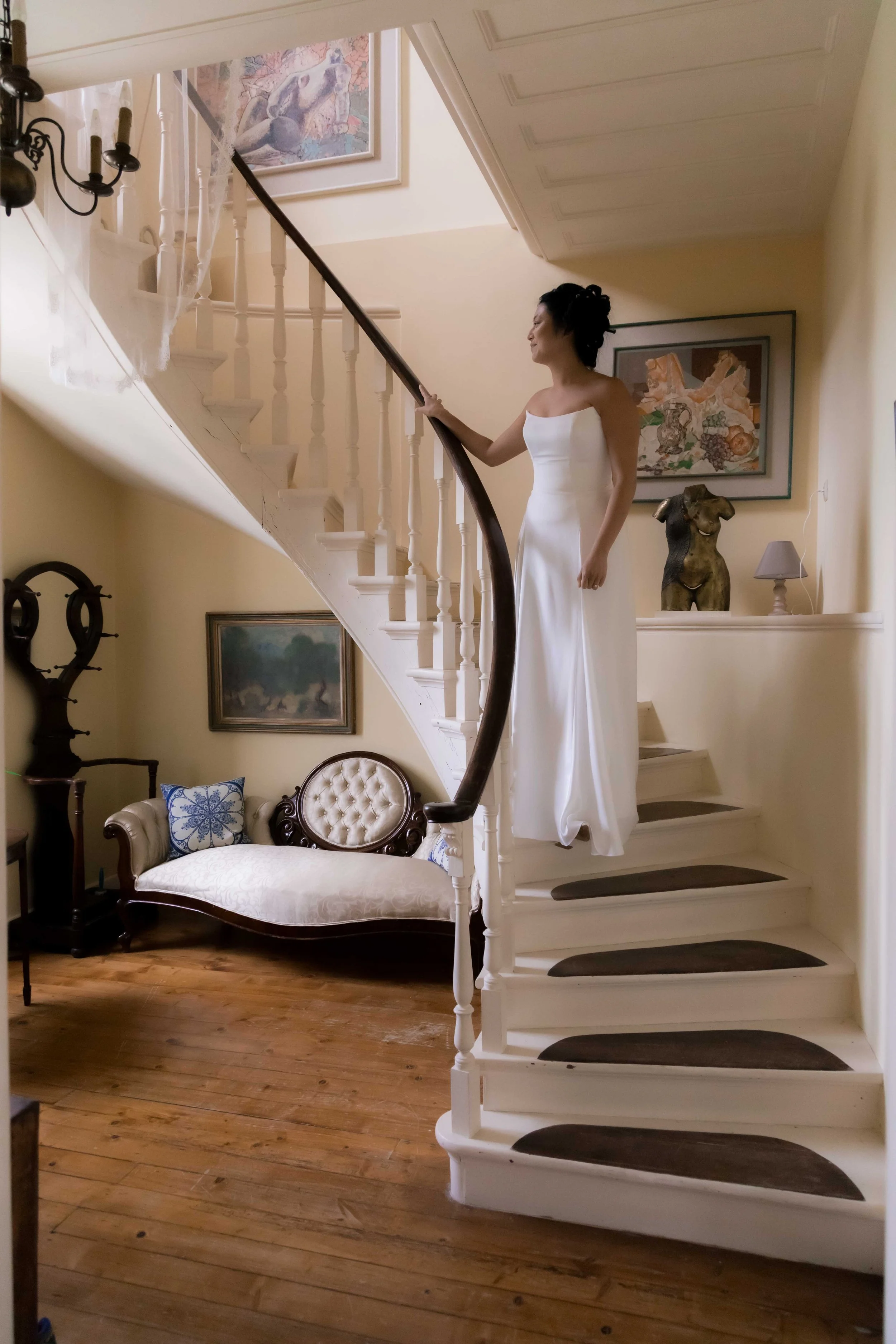 A woman in a white dress standing on the staircase landing of a home, holding the railing and looking to the side. The staircase curves upward with dark treads and white risers. The room has framed artwork, a vintage bench with pillows, a sculpture o