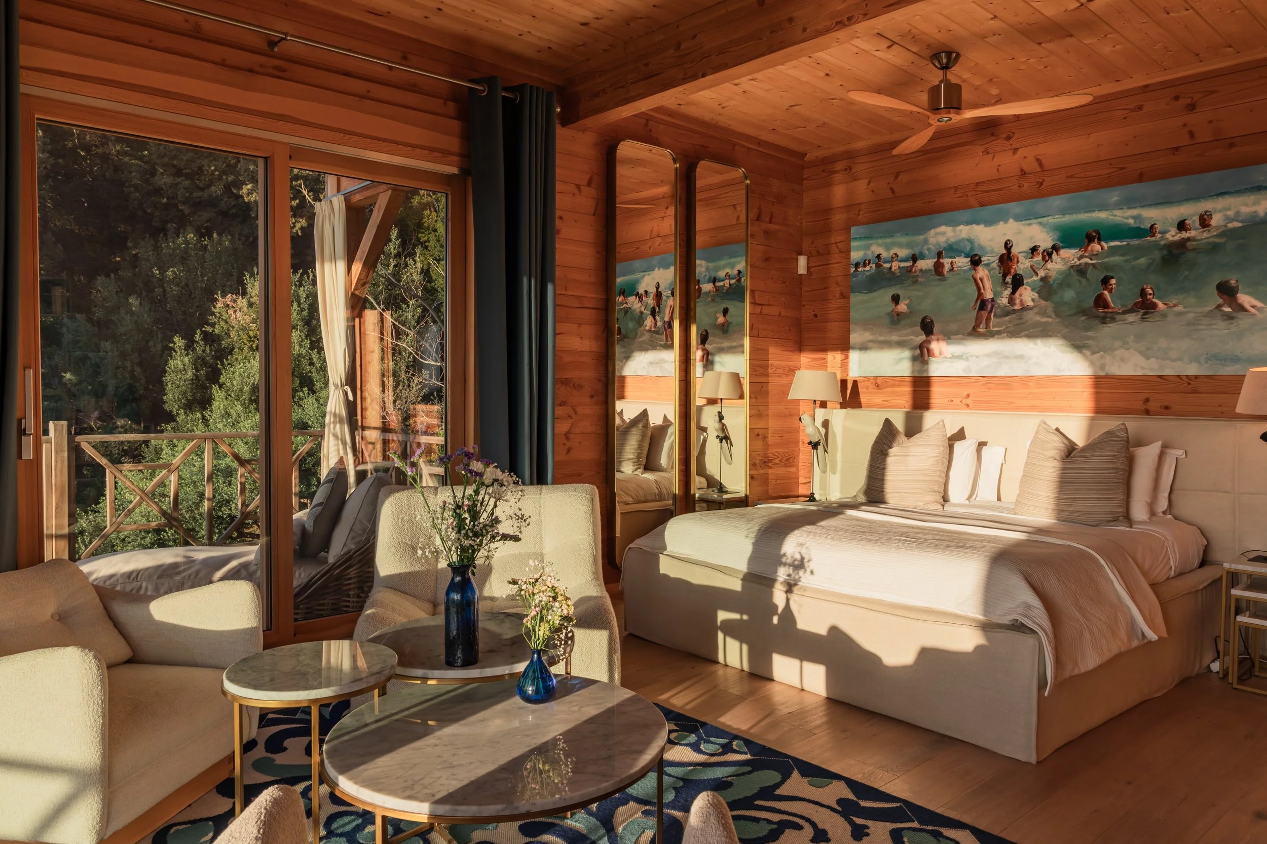 A cozy bedroom with wooden walls and ceiling, featuring a large bed with cream-colored linens, two mirrors, and wall art of people at the beach. There's a sitting area with a sofa and round coffee tables near a large sliding glass door leading to a b