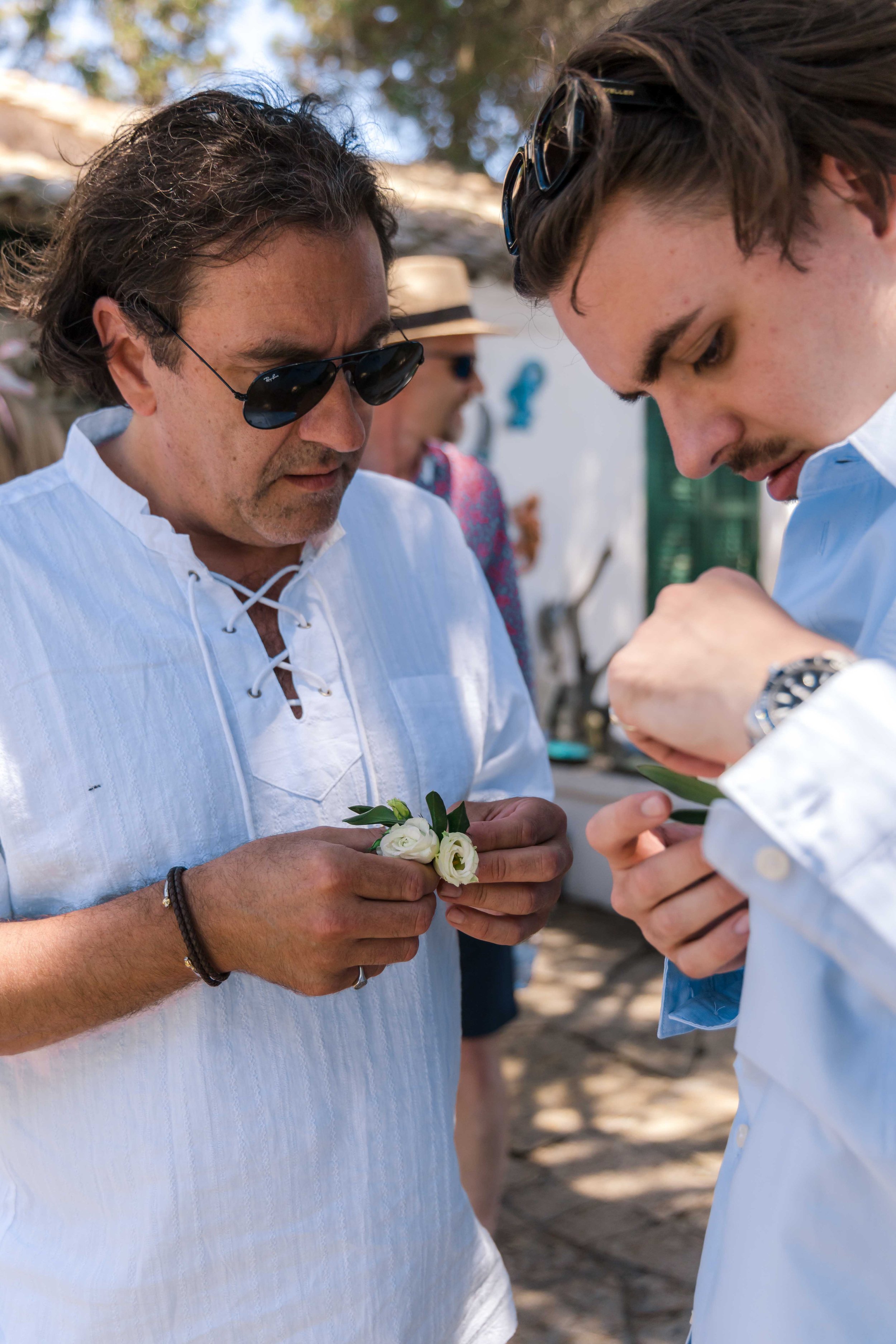 Two men wearing white shirts handling small flower arrangements outdoors at a sunny event, with other people in the background.