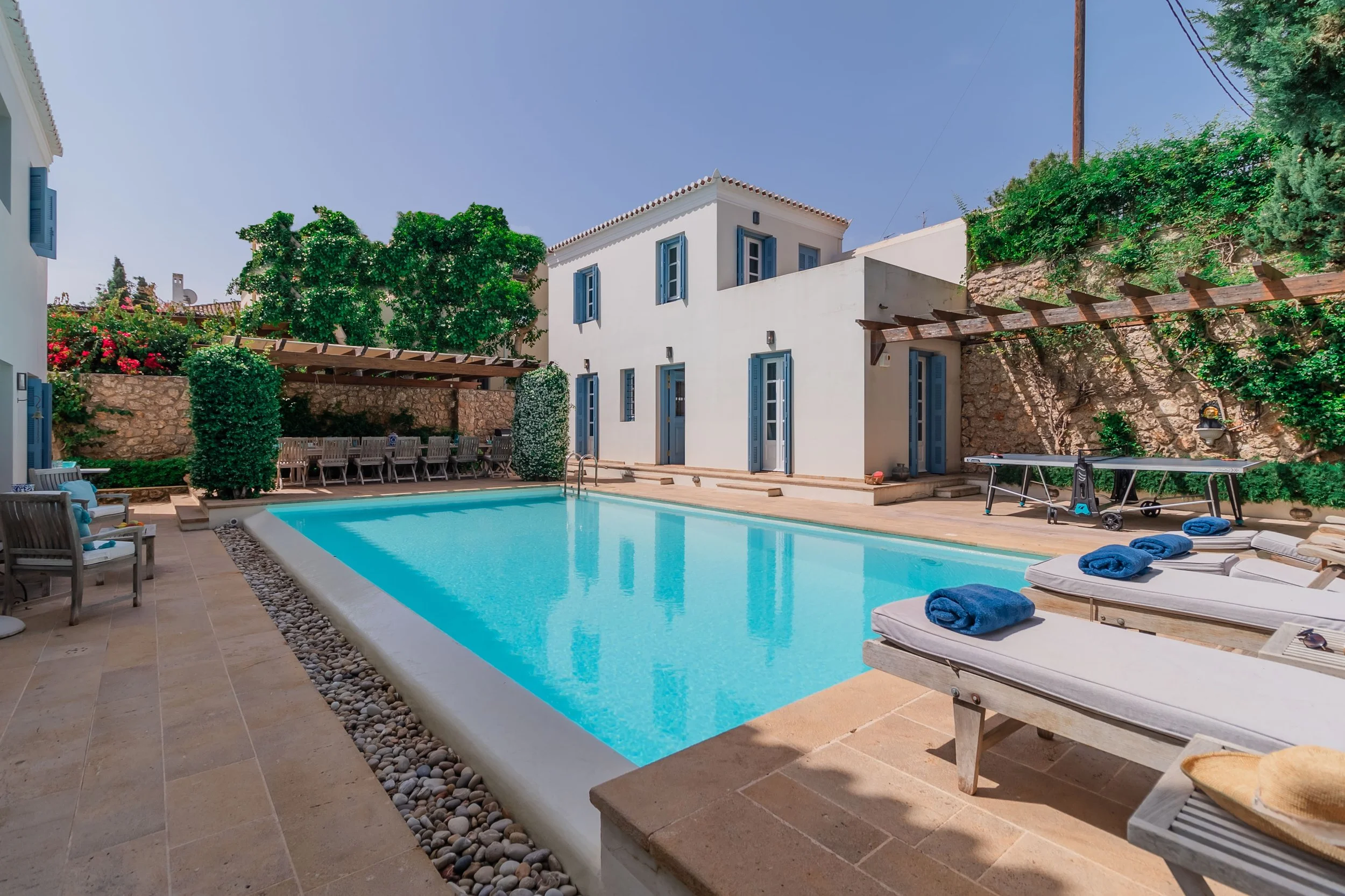 A luxurious backyard with a rectangular swimming pool, sun loungers with blue towels, a shaded dining area with a long table and chairs, a ping pong table, white house with blue window shutters, stone and wooden fencing, greenery, and clear blue sky.