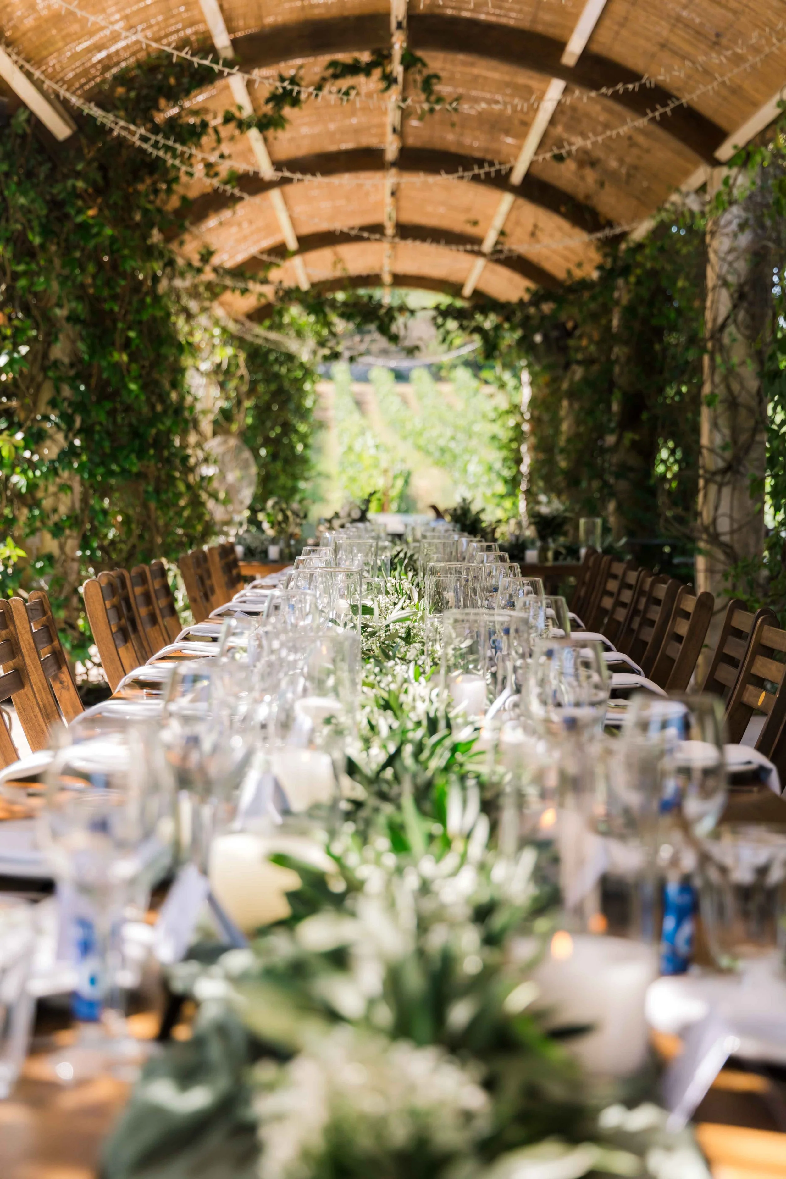 Long table set for a formal outdoor event under a wooden covered patio, decorated with white flowers and greenery, with sunlight filtering through the surrounding trees.