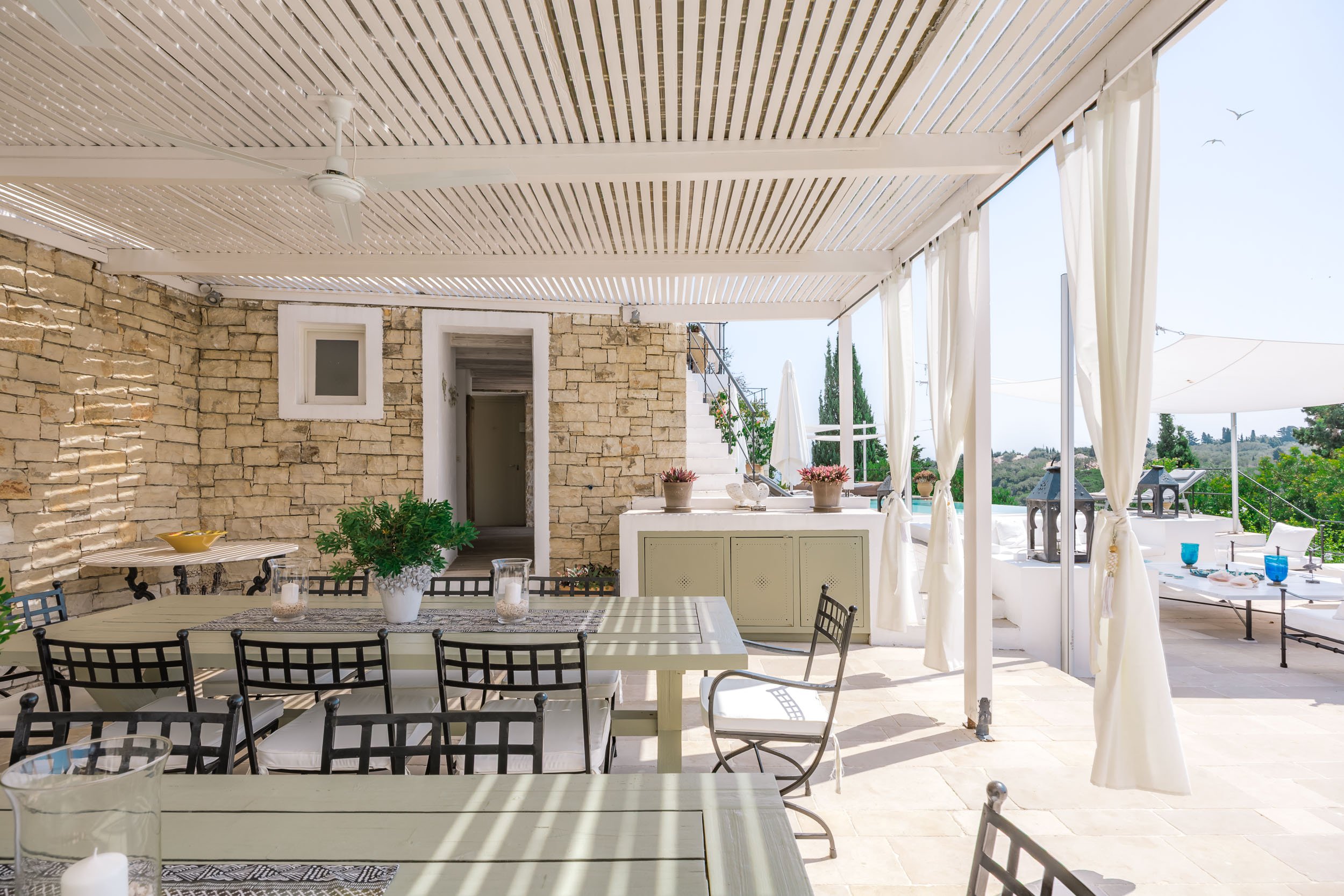 Outdoor patio area with dining table, chairs, potted plants, stone wall, and view of trees and sky in the background.
