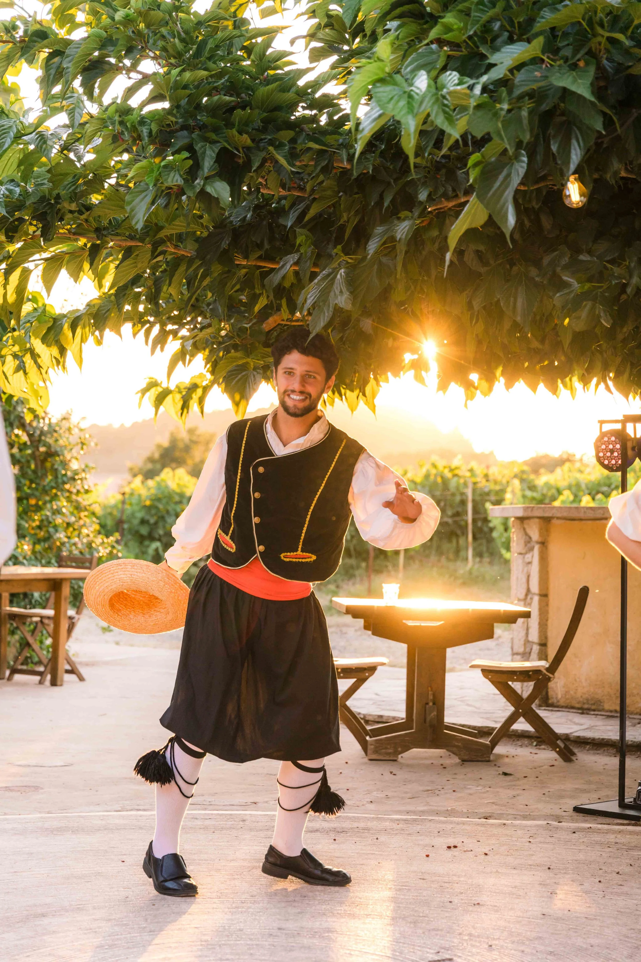 A man wearing traditional Greek clothing, including a black kilt, white shirt, and black vest, holding a straw hat and smiling outdoors at sunset under a large tree with green leaves.
