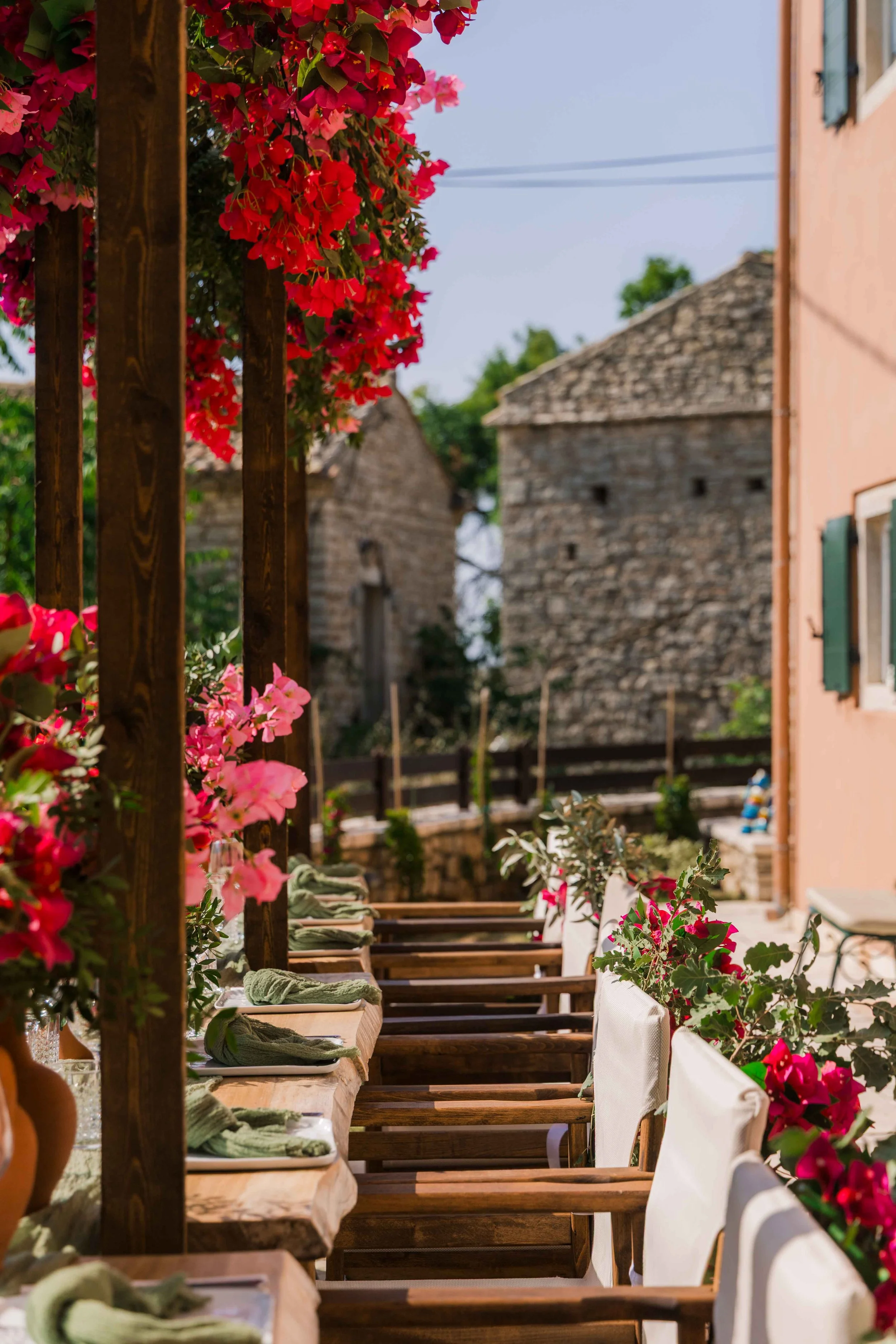Outdoor dining setup with wooden tables, green napkins, and white chairs, decorated with pink and red flowers along a rustic fence, with stone buildings and trees in the background.