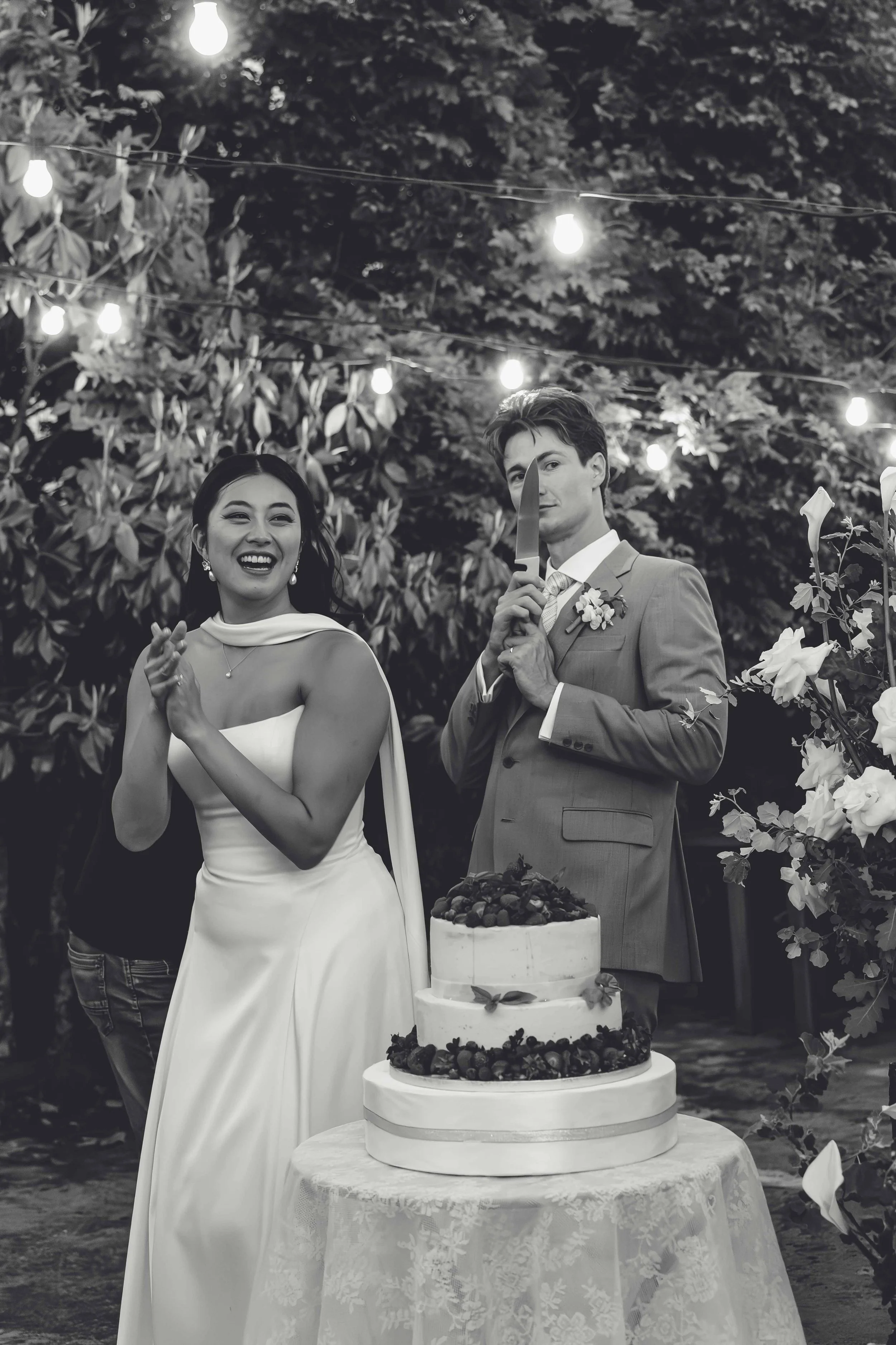 Black and white photo of a wedding celebration outdoors featuring a bride and groom. The bride is laughing and clapping, wearing a white wedding dress, while the groom, dressed in a suit, is holding a large knife, as if about to cut the wedding cake.