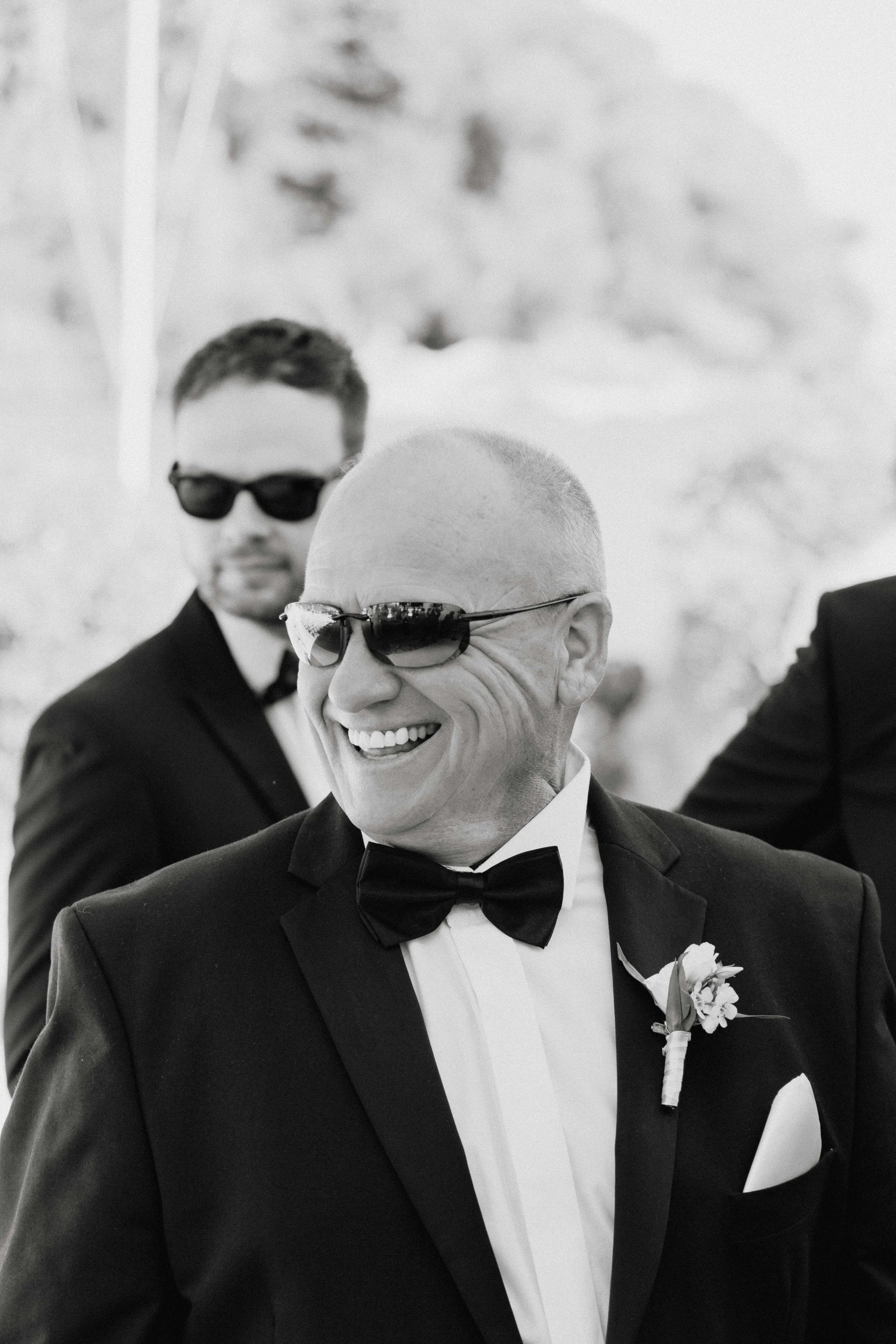 A black-and-white photo of smiling man in tuxedo with bow tie and sunglasses, with another man in tuxedo and sunglasses in the background, outdoors.