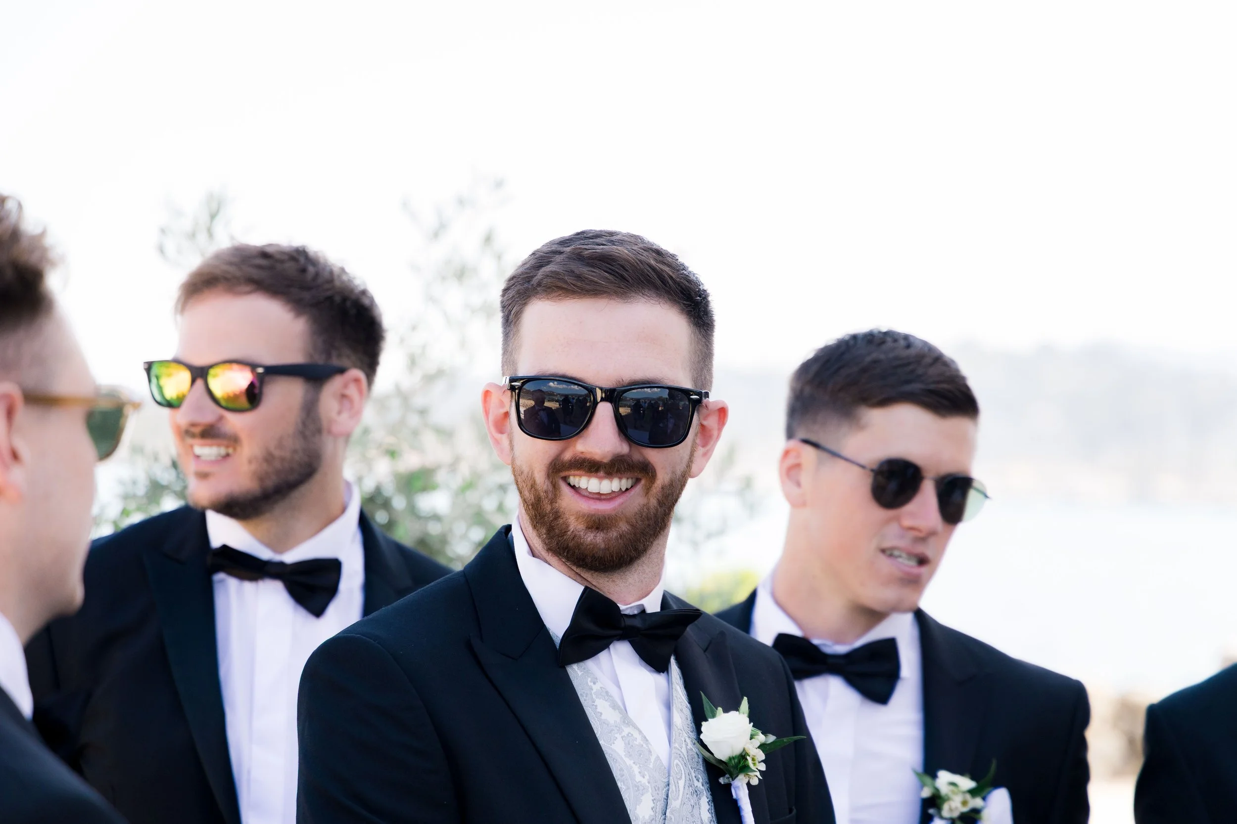 A group of men dressed in tuxedos and sunglasses, smiling and engaging in conversation outdoors.