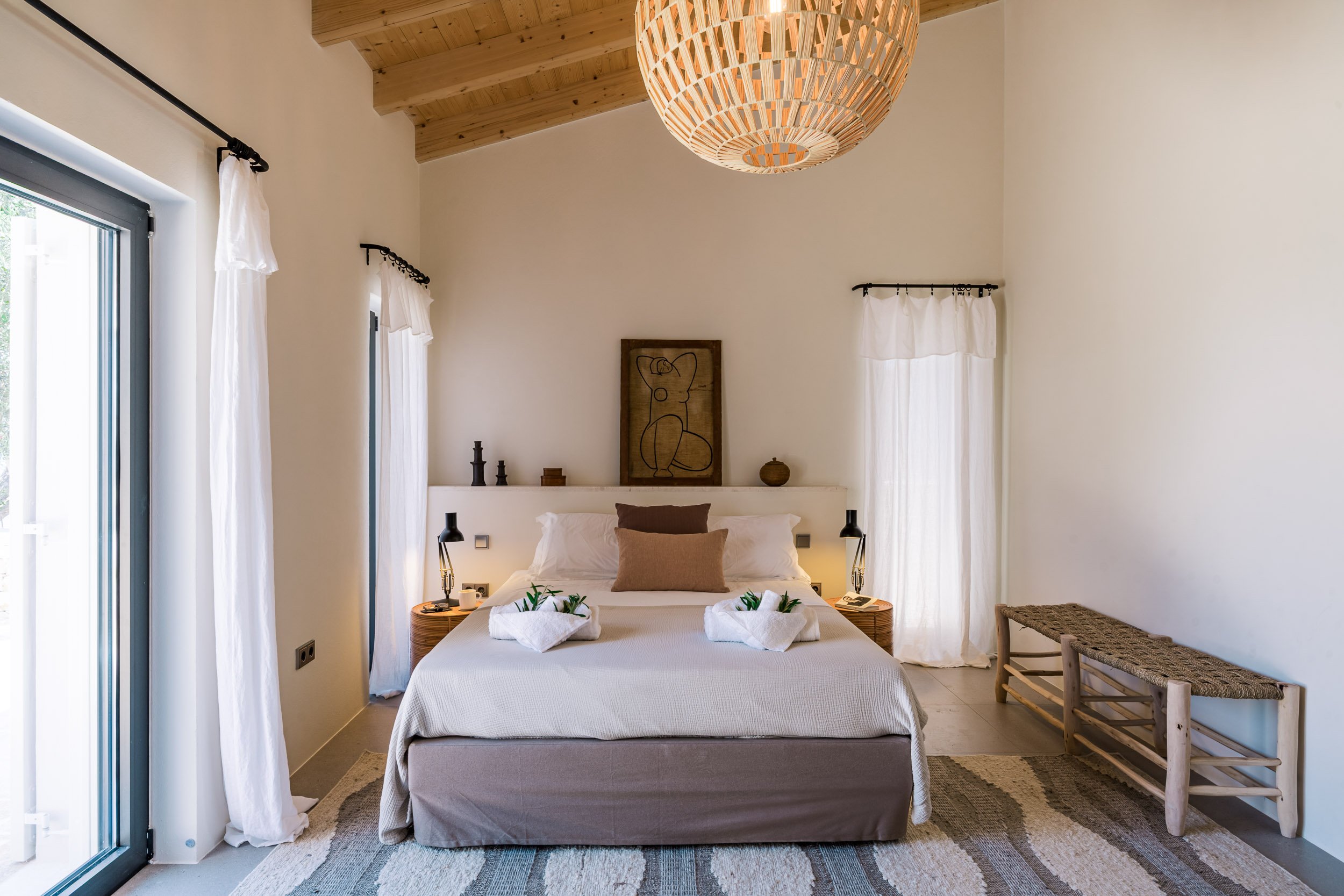 A bedroom with a neutral color scheme, featuring a bed with pillows and towels with greenery, two nightstands with lamps, a wooden bench, and natural light coming through white curtains, with a framed abstract drawing above the headboard.