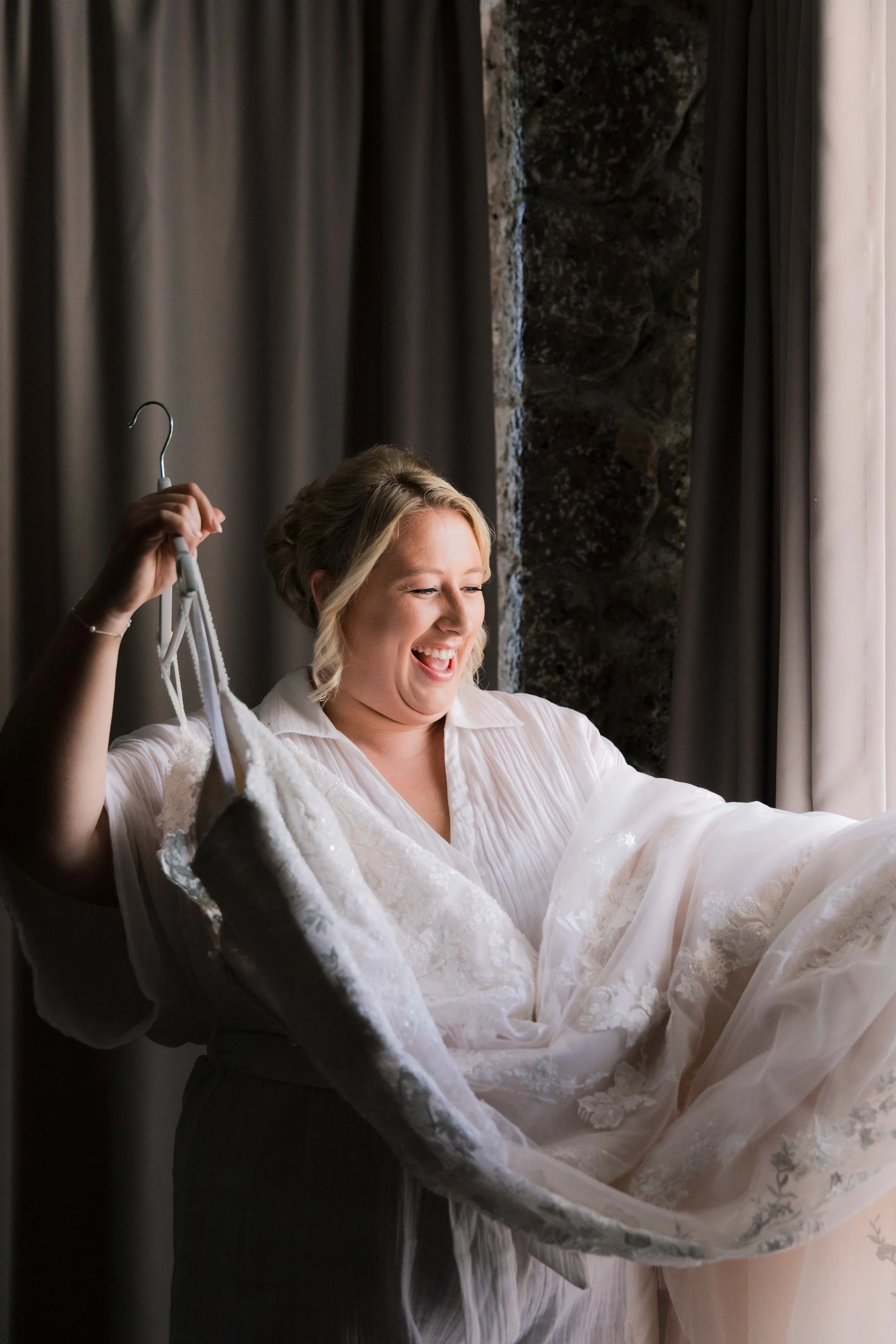 A woman in a white robe holding a wedding dress with lace details, smiling joyfully as she looks at it.