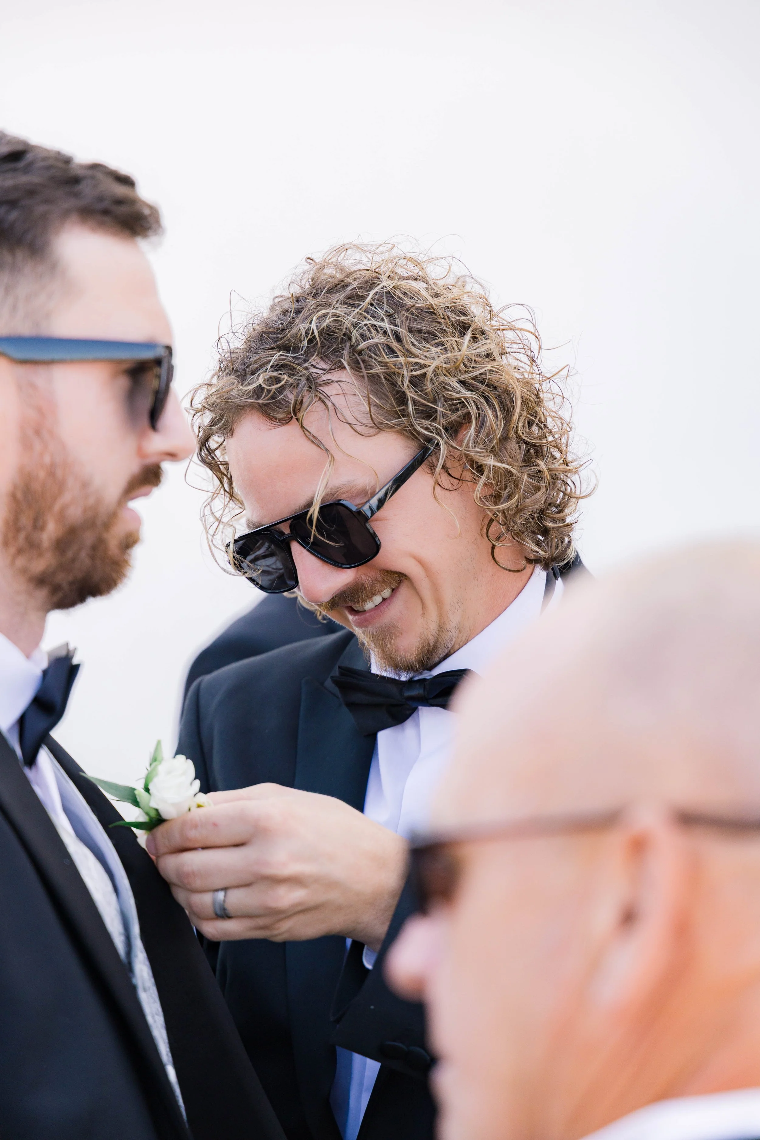Men in tuxedos and bow ties wearing sunglasses, smiling and talking at a wedding event.