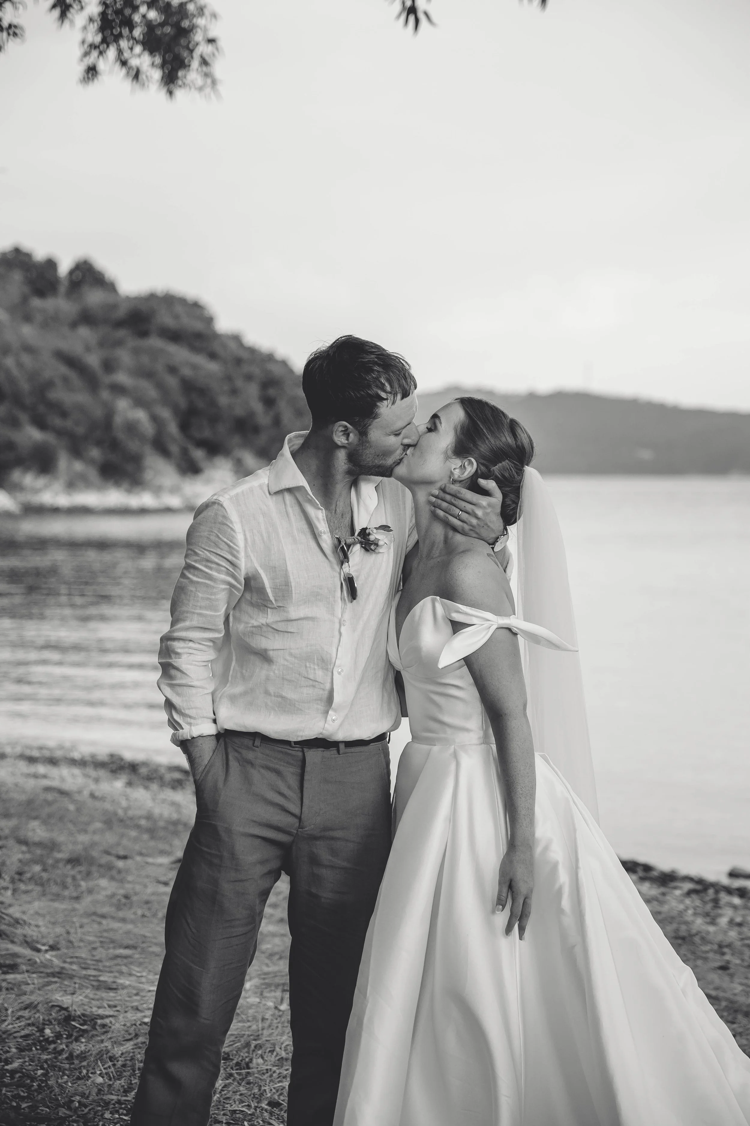 A black and white photo of a man and woman kissing by a lakeside, wedding attire, woman with veil, man in dress shirt and trousers.