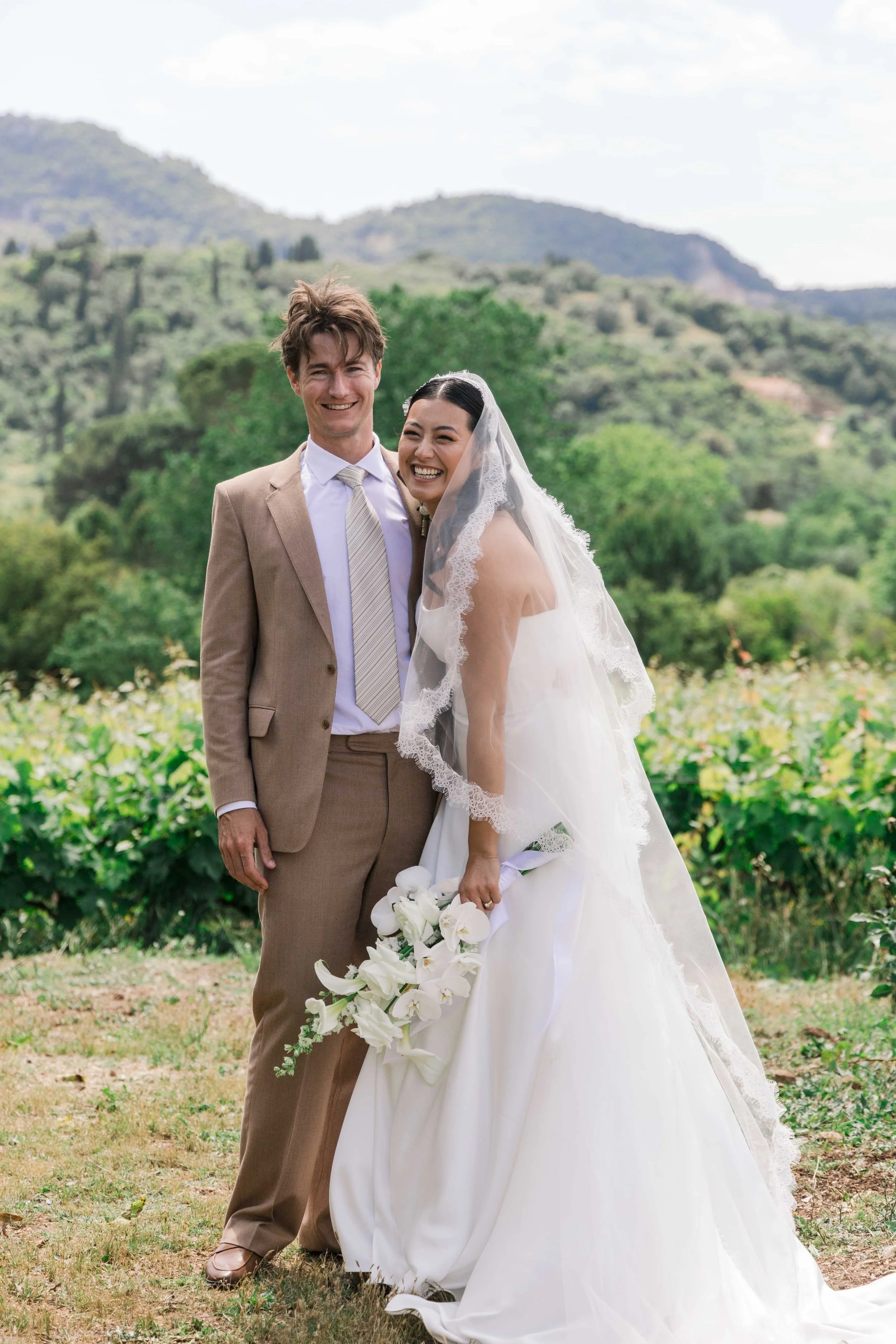 A happy bride and groom standing outdoors on a wedding day, smiling, with green hills and trees in the background. The bride is holding a white bouquet and wearing a white wedding dress with a lace veil, while the groom is dressed in a beige suit wit