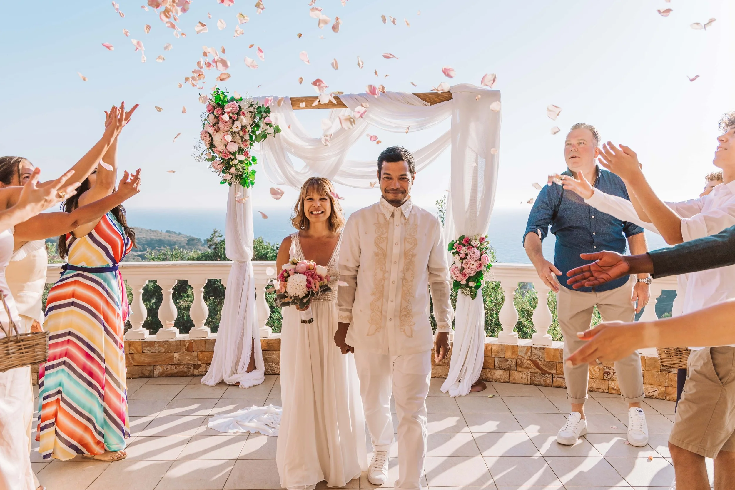 A couple in wedding attire walking down the aisle under a floral arch, surrounded by friends throwing flower petals on a sunny outdoor terrace with ocean in the background.