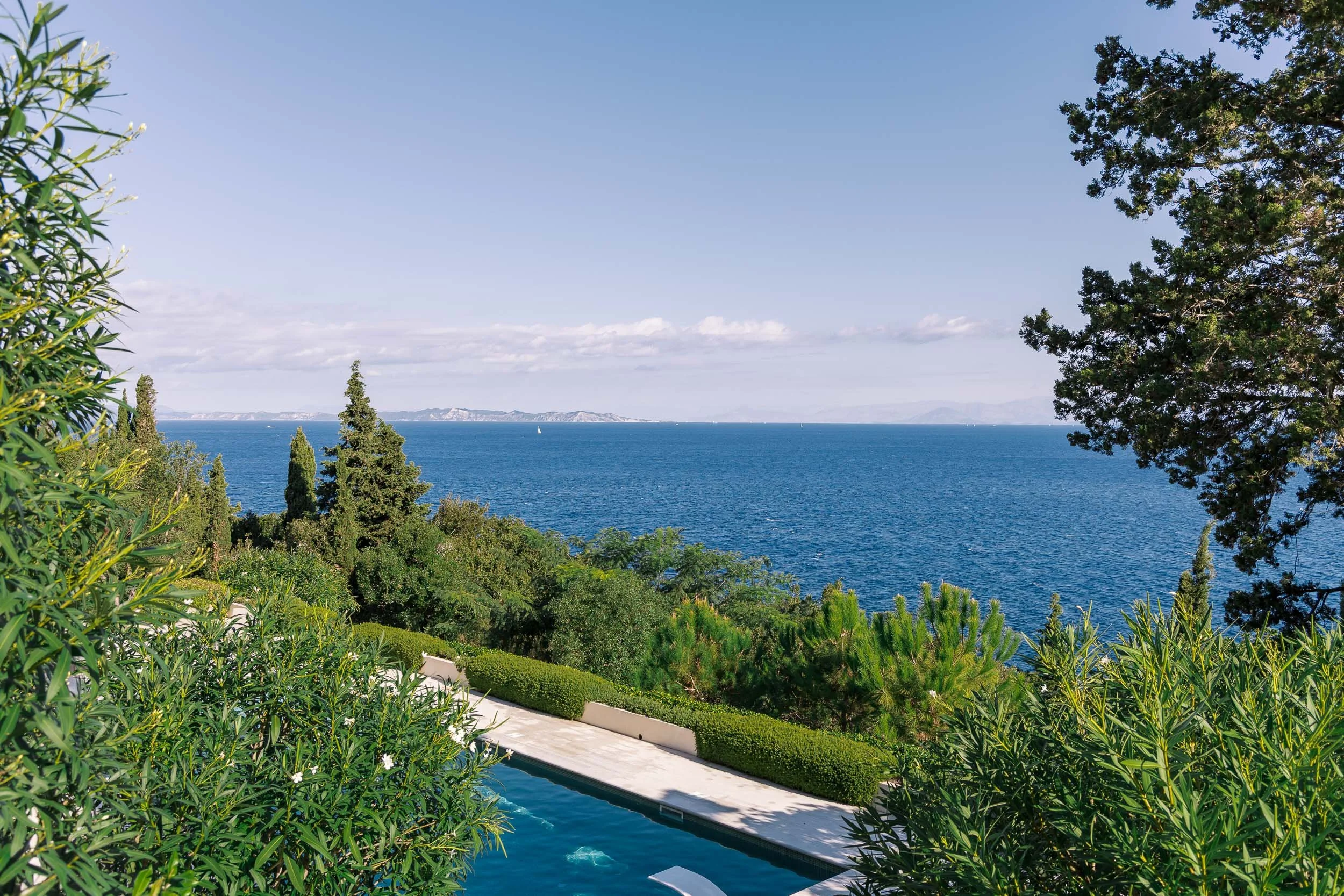 View of ocean through trees and greenery, with a swimming pool in the foreground and mountains in the distance.