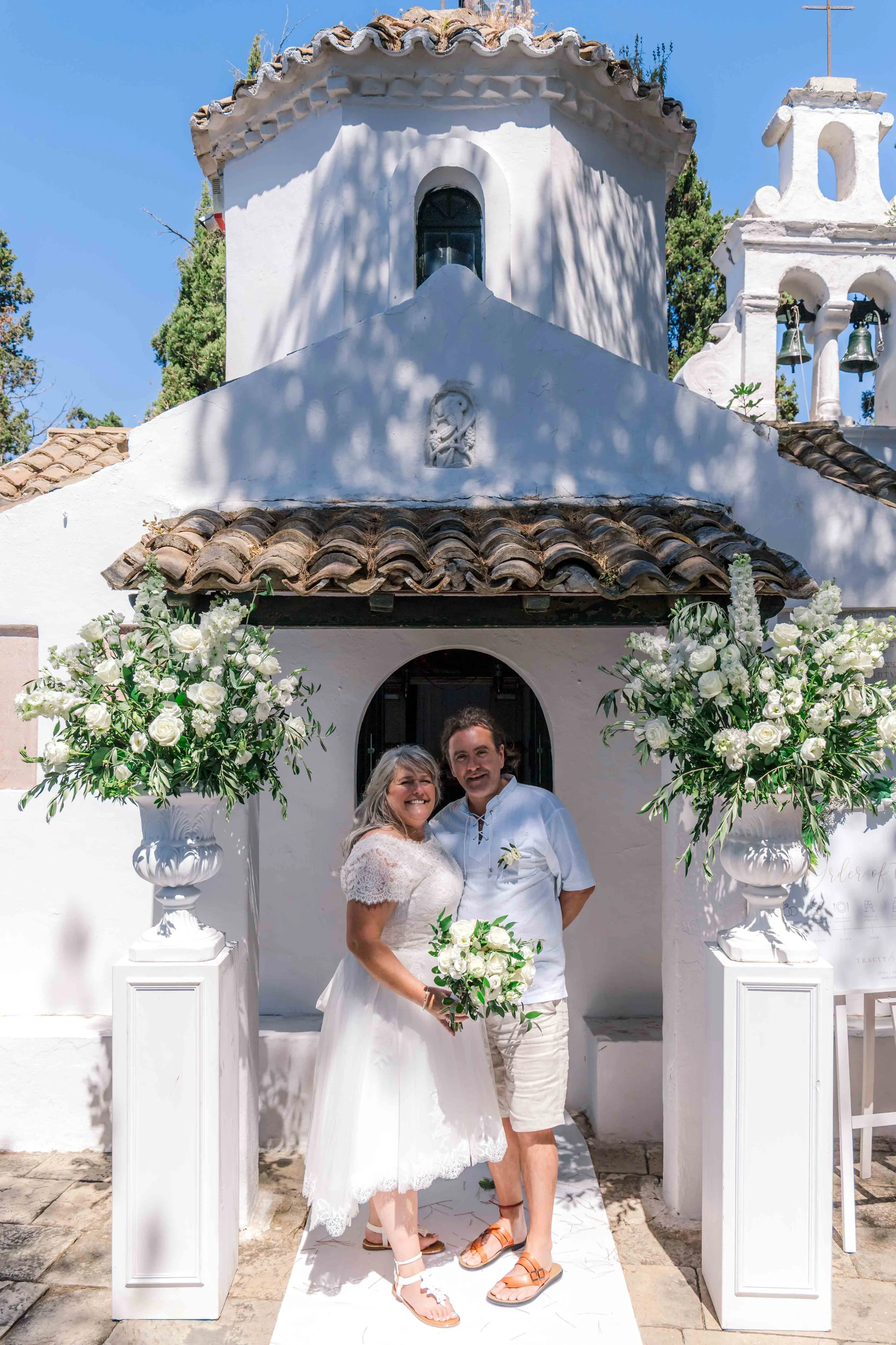 A newly married couple is standing in front of a white church with two large floral arrangements on white pedestals. The bride is wearing a white lace dress and holding a bouquet, and the groom is dressed in casual white attire. The church has a smal