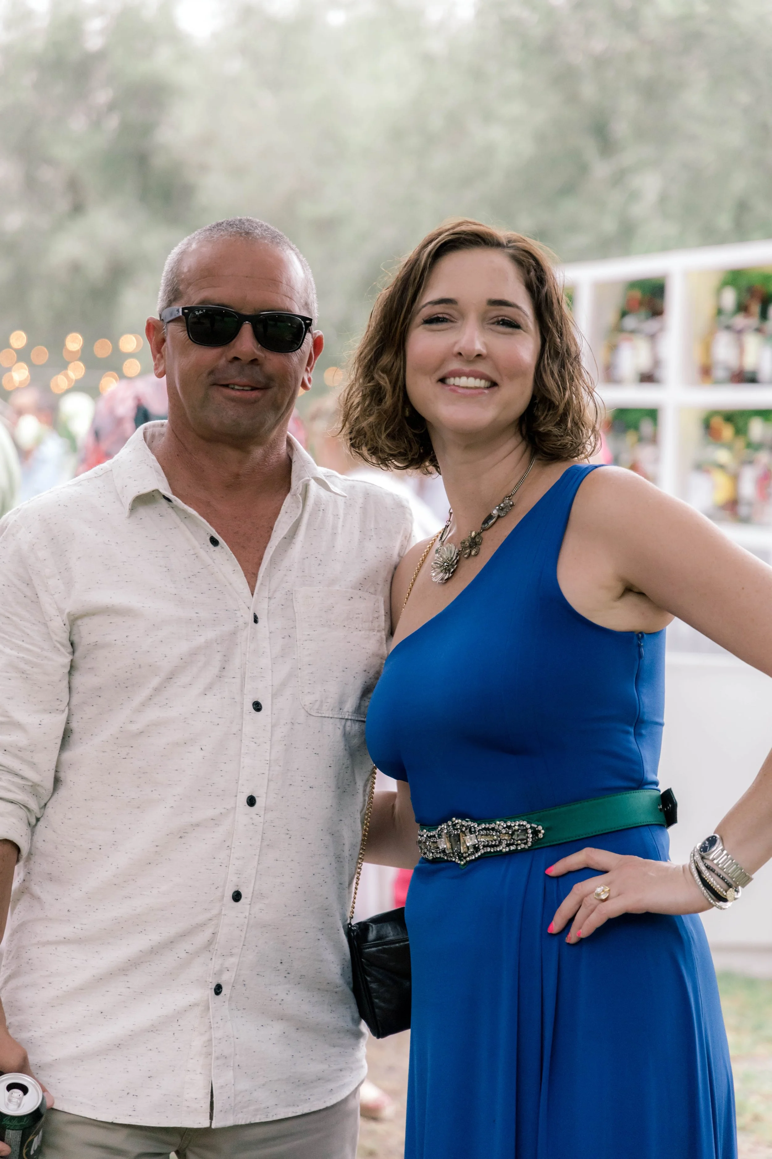 A man wearing sunglasses and a light-colored shirt standing next to a woman in a blue dress with jewelry, at an outdoor event or festival.