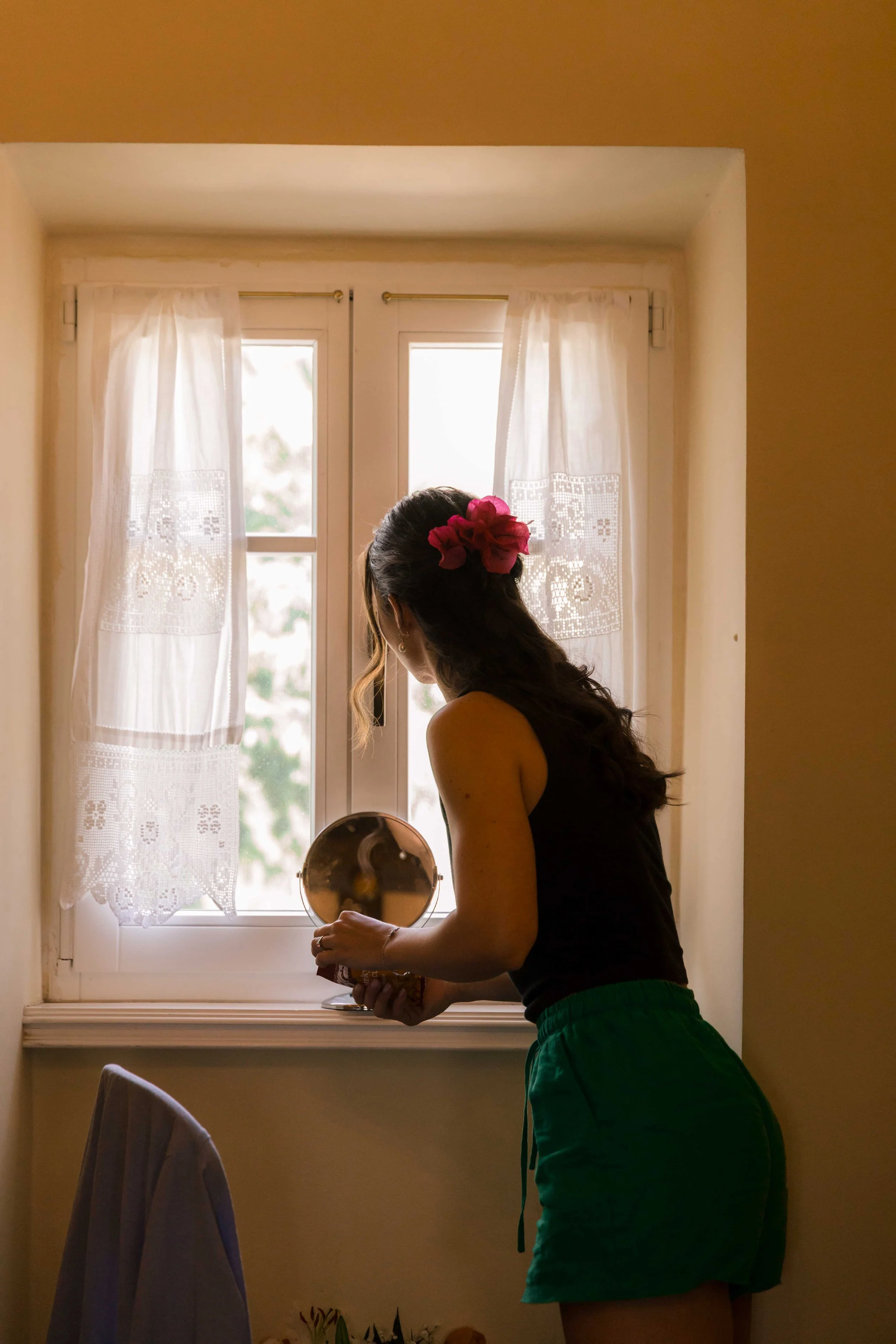 A woman with long dark hair, adorned with a pink flower, looking into a small round mirror near a window with white lace curtains, dressed in a black top and green shorts.