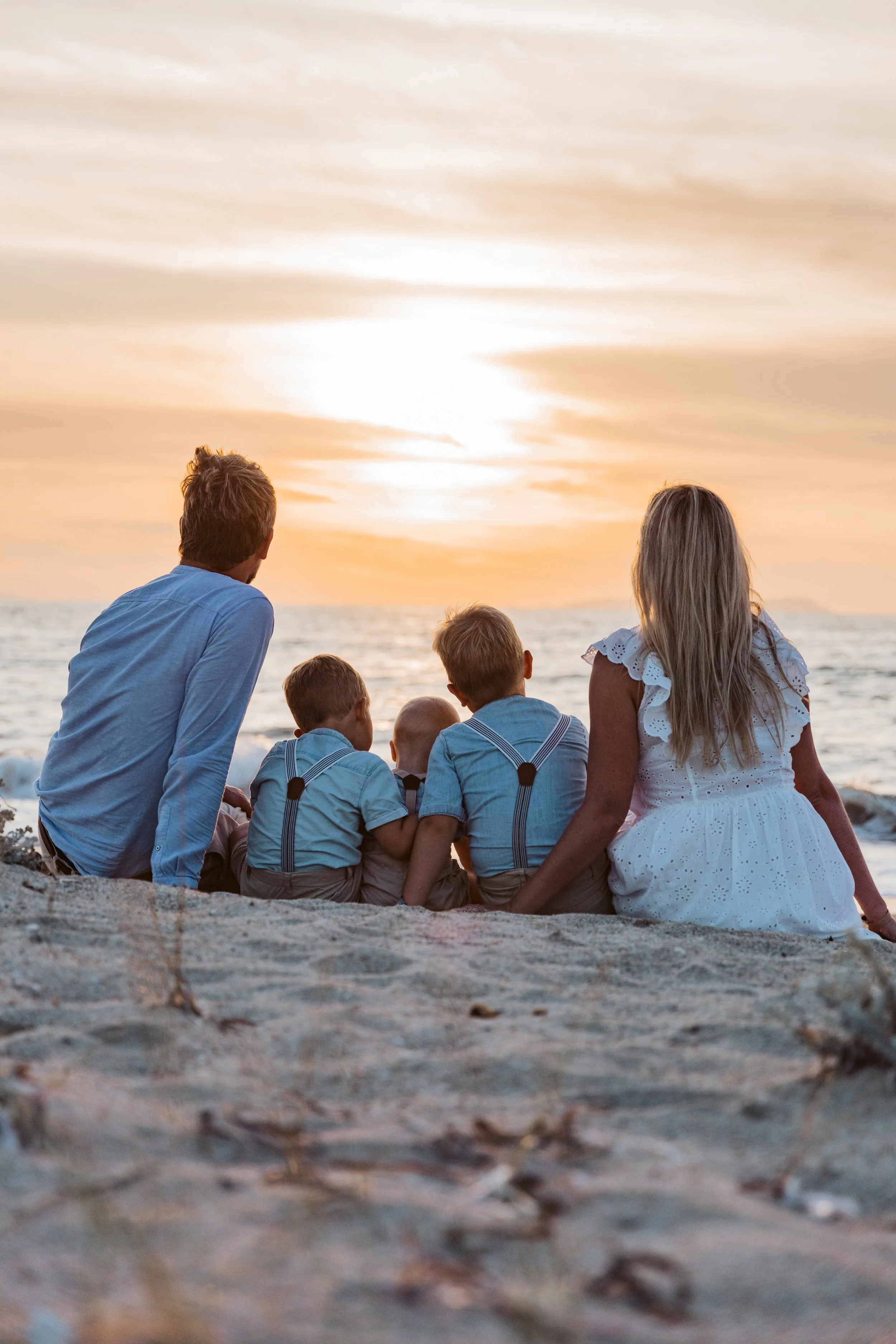 Family of five sitting on the beach at sunset, facing the ocean.