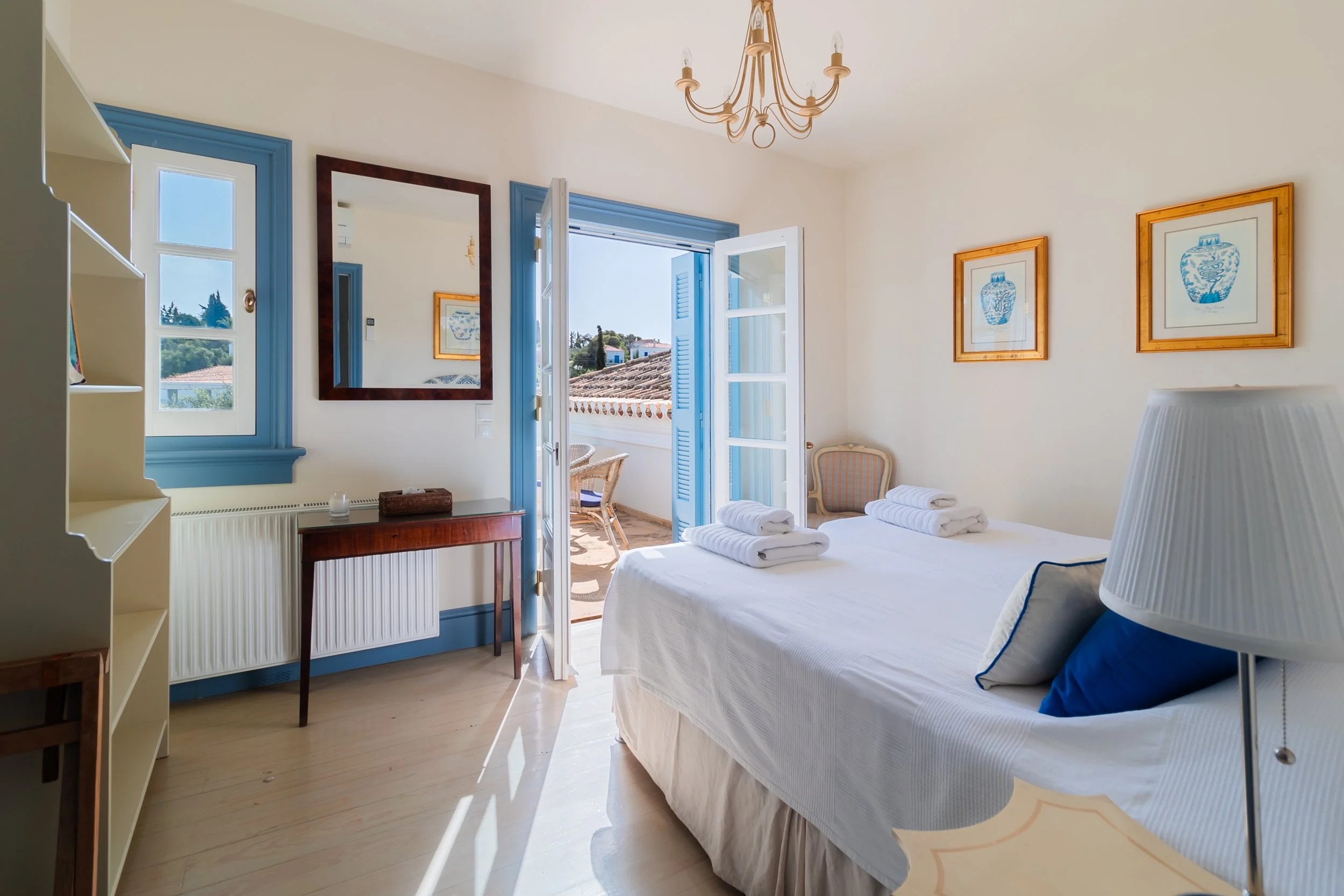A bright hotel bedroom with a balcony, white walls, blue window frames, a mirror and framed art, a bed with white linens and blue pillows, and a small sitting area outside on the balcony.
