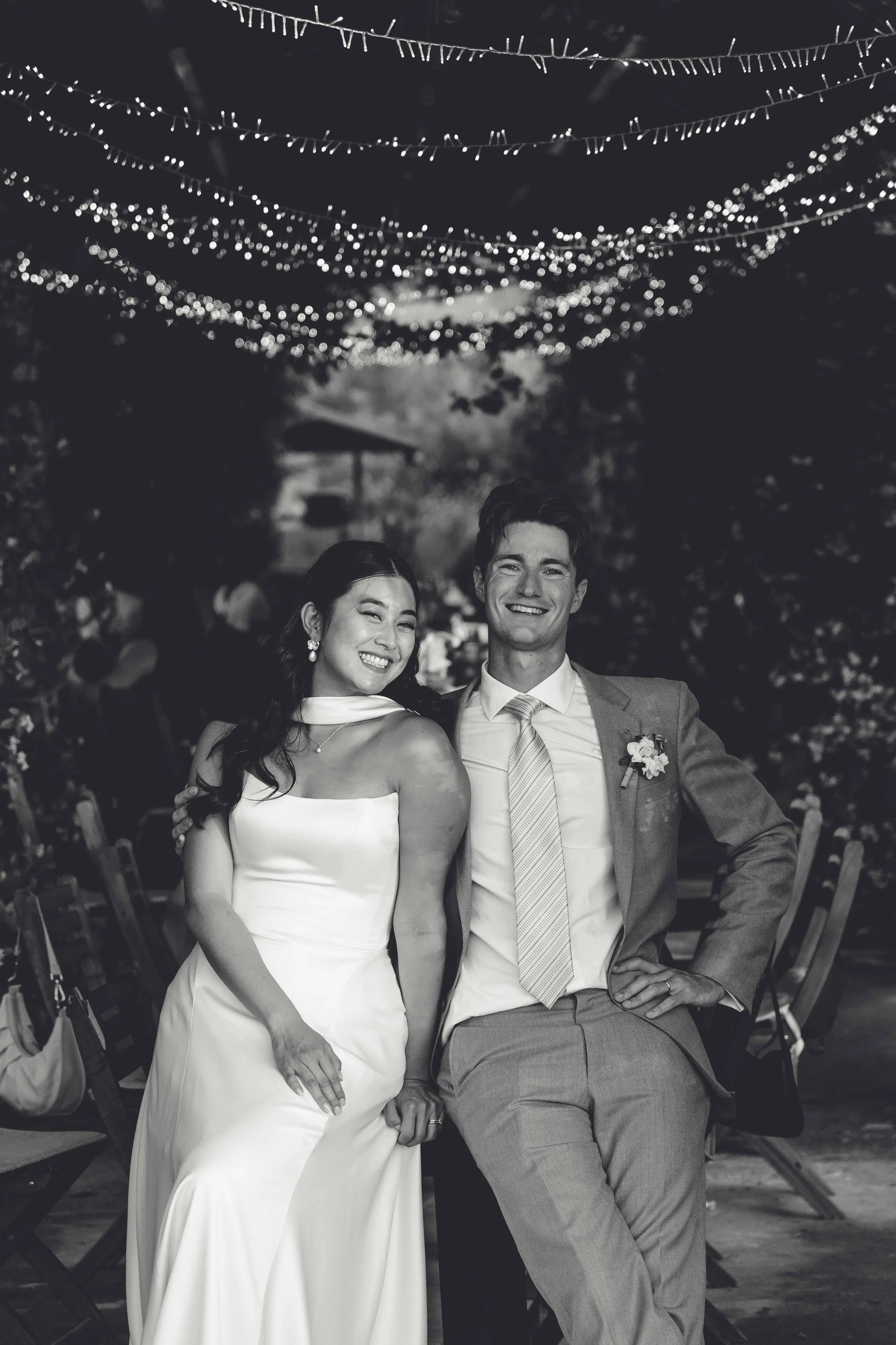 A black-and-white photo of a newlywed couple smiling, holding hands, sitting at a wedding reception under string lights, with guests and trees in the background.