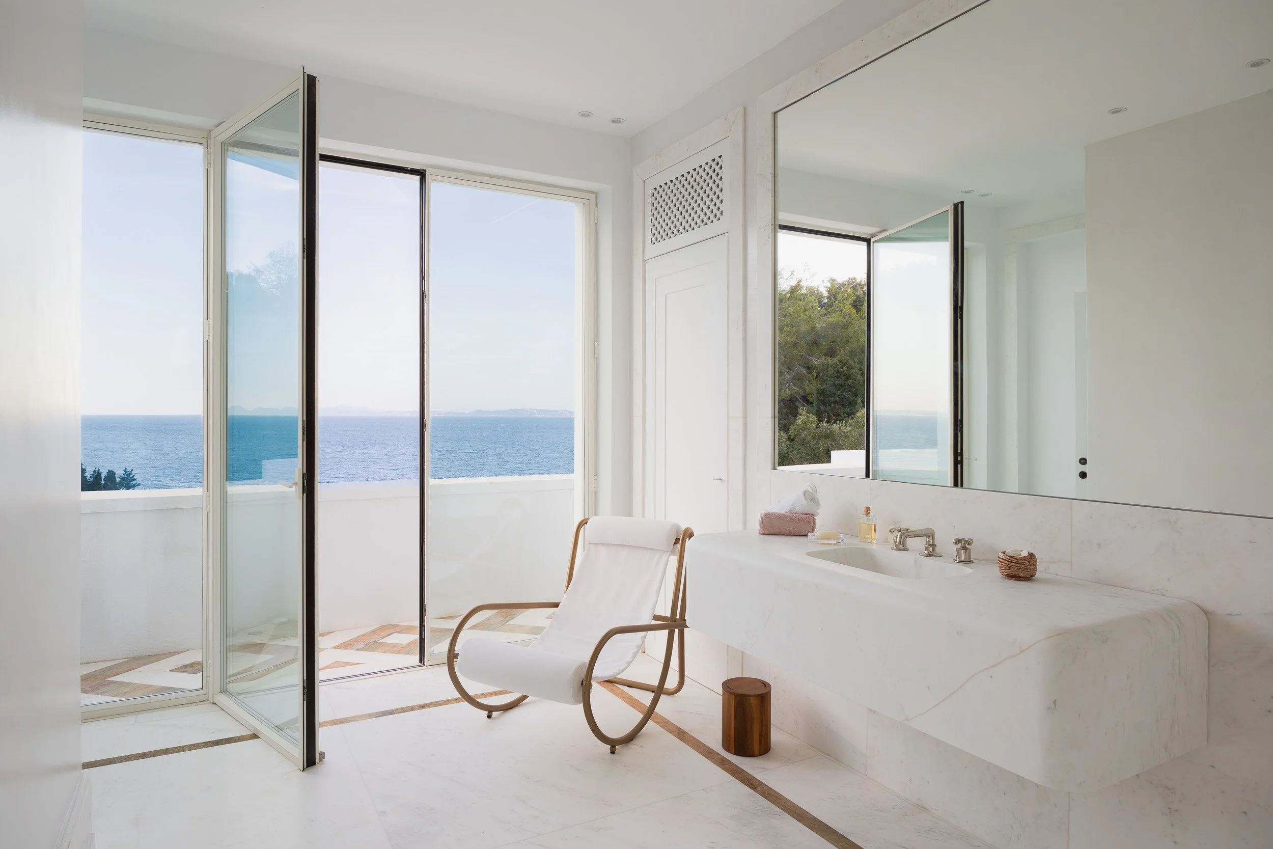 Bright bathroom with large windows and ocean view, modern white marble sink, white chair, small wooden stool, and minimal decor.