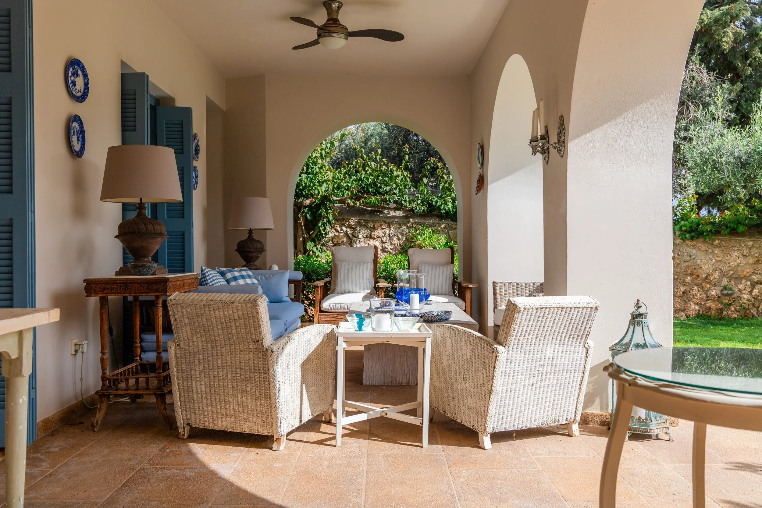 Covered outdoor patio with wicker chairs, a wooden side table with lamps, and a small round table with a glass top, overlooking a garden with trees and stone wall.