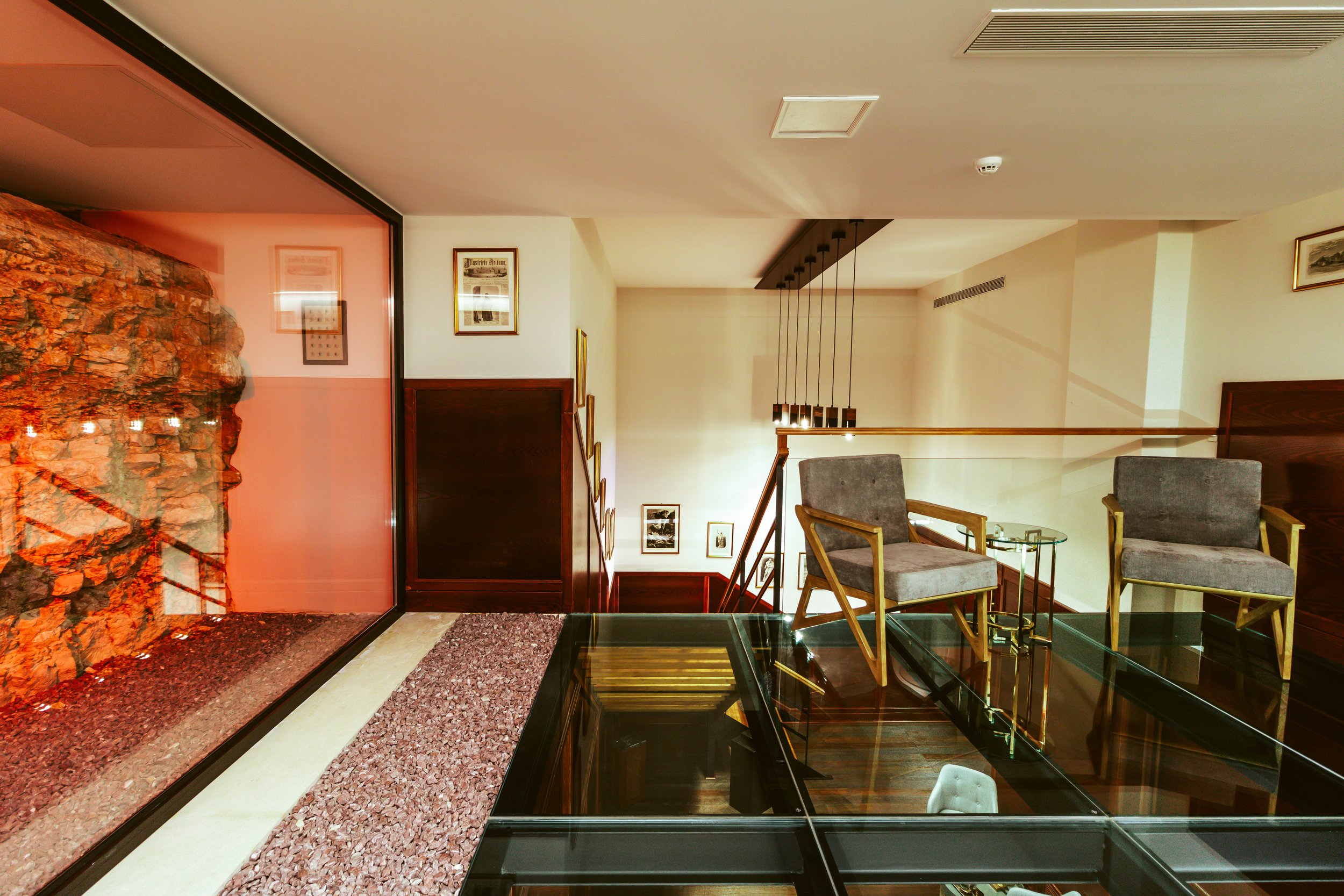 Interior of a modern lounge area with two gray armchairs, a glass table, framed pictures on the wall, and a glass display case in the foreground. A large glass partition with a red hue and exposed brick wall is visible on the left.