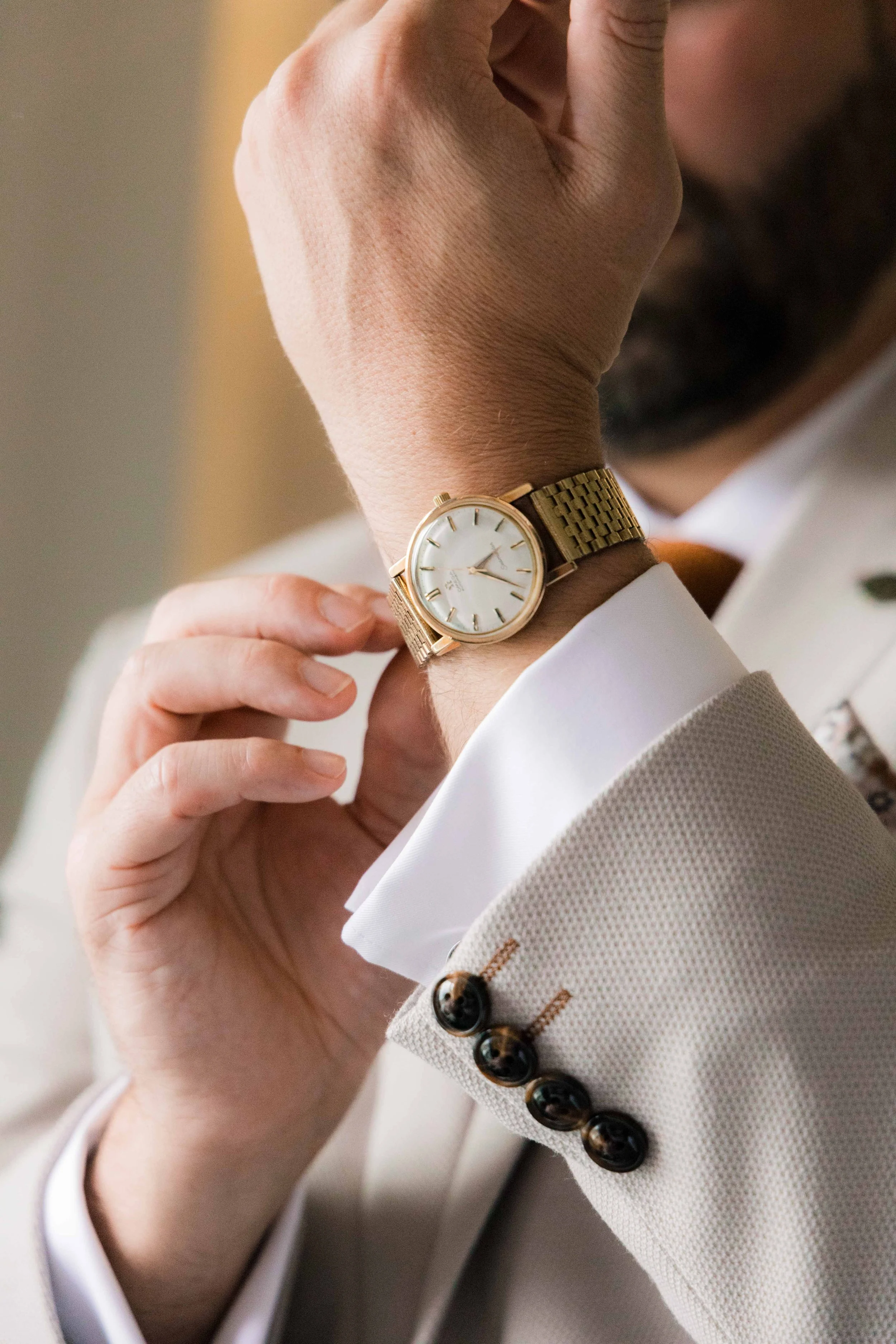 Close-up of a man wearing a beige suit with dark buttons, a white dress shirt, and a gold wristwatch with a white face. The man is adjusting his watch with his hand near his face.