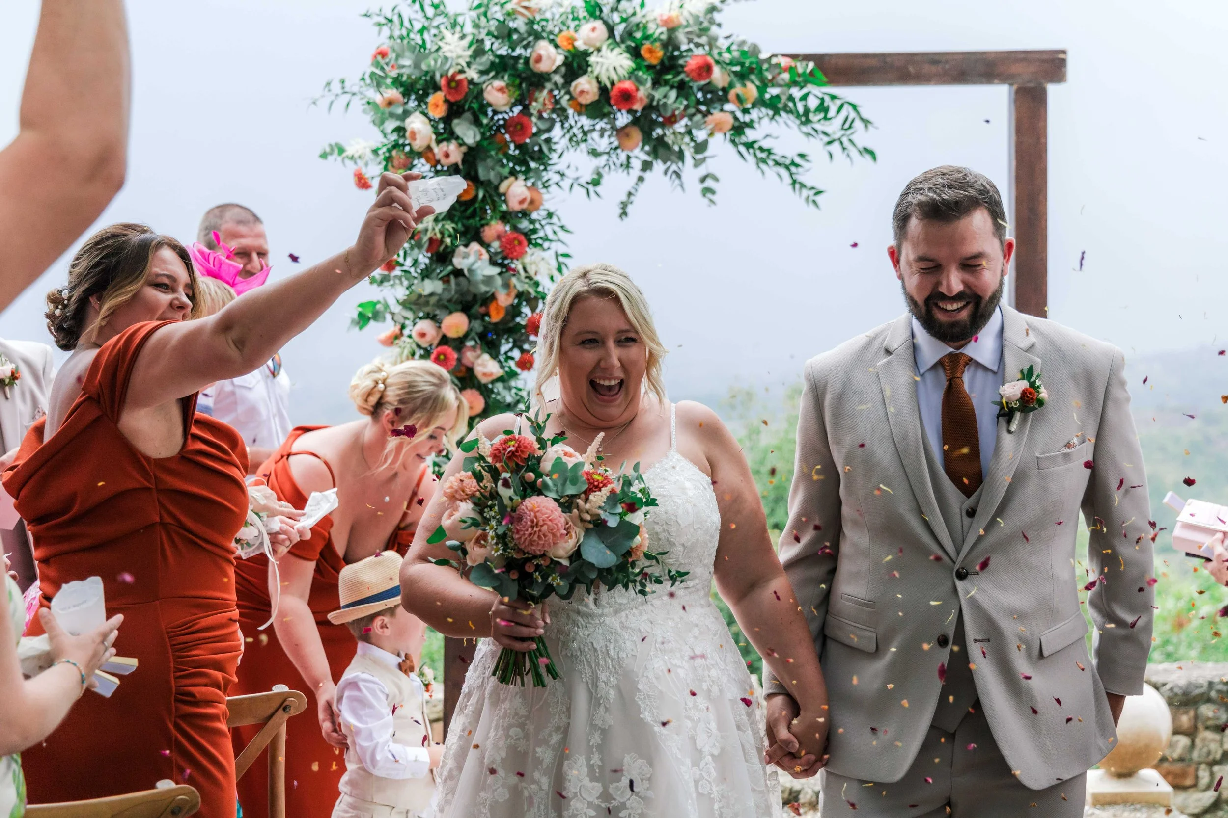 Bride and groom holding hands and smiling at their wedding celebration, surrounded by floral arrangements and guests throwing confetti.