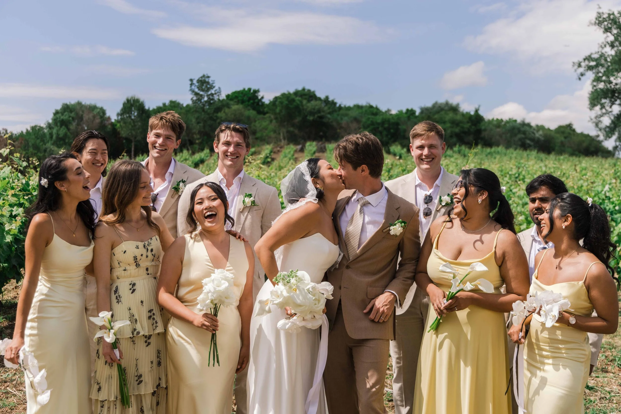 A wedding with a bride and groom kissing, surrounded by bridesmaids and groomsmen outdoors on a sunny day, with greenery in the background.