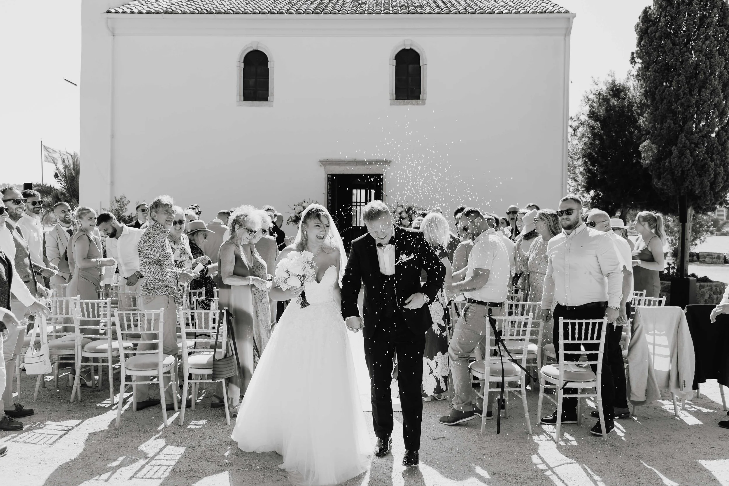 A black and white photo of a wedding reception outside a church with the bride and groom walking hand in hand through guests, who are cheering and throwing confetti.