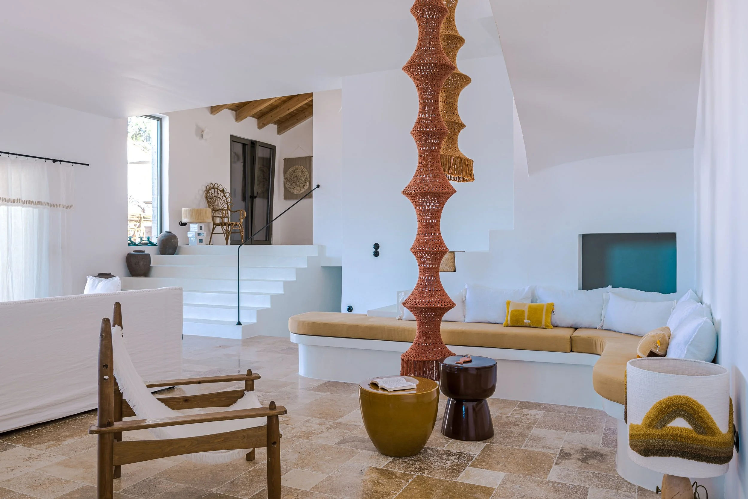 Modern living room with white walls, a curved beige sectional sofa with white and yellow pillows, a wooden chair with white upholstery, a multicolored ceramic side table, and a tall sculptural wooden divider. There are white stairs leading to a small