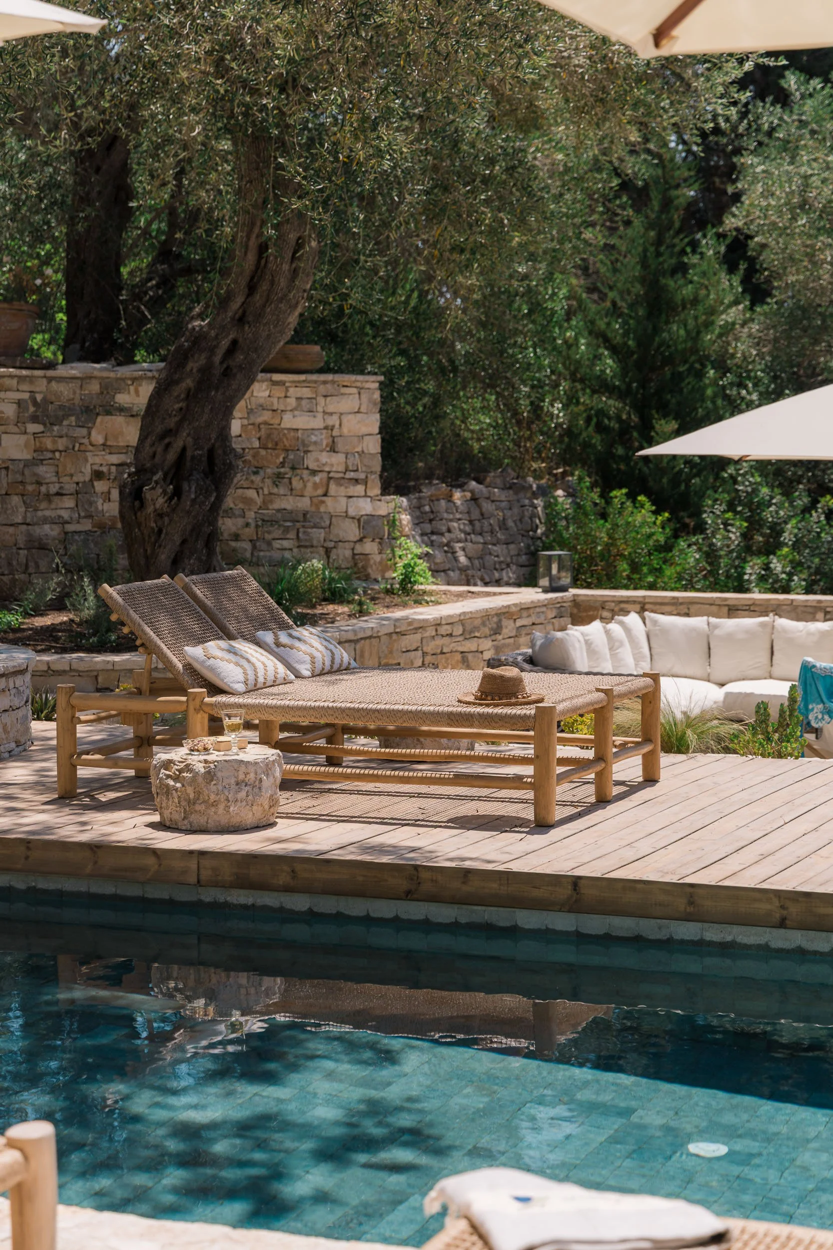 A backyard poolside scene with a wooden lounge chair, a straw hat, a glass of beverage, a stone side table, and cushioned seating, surrounded by lush trees and stone walls.