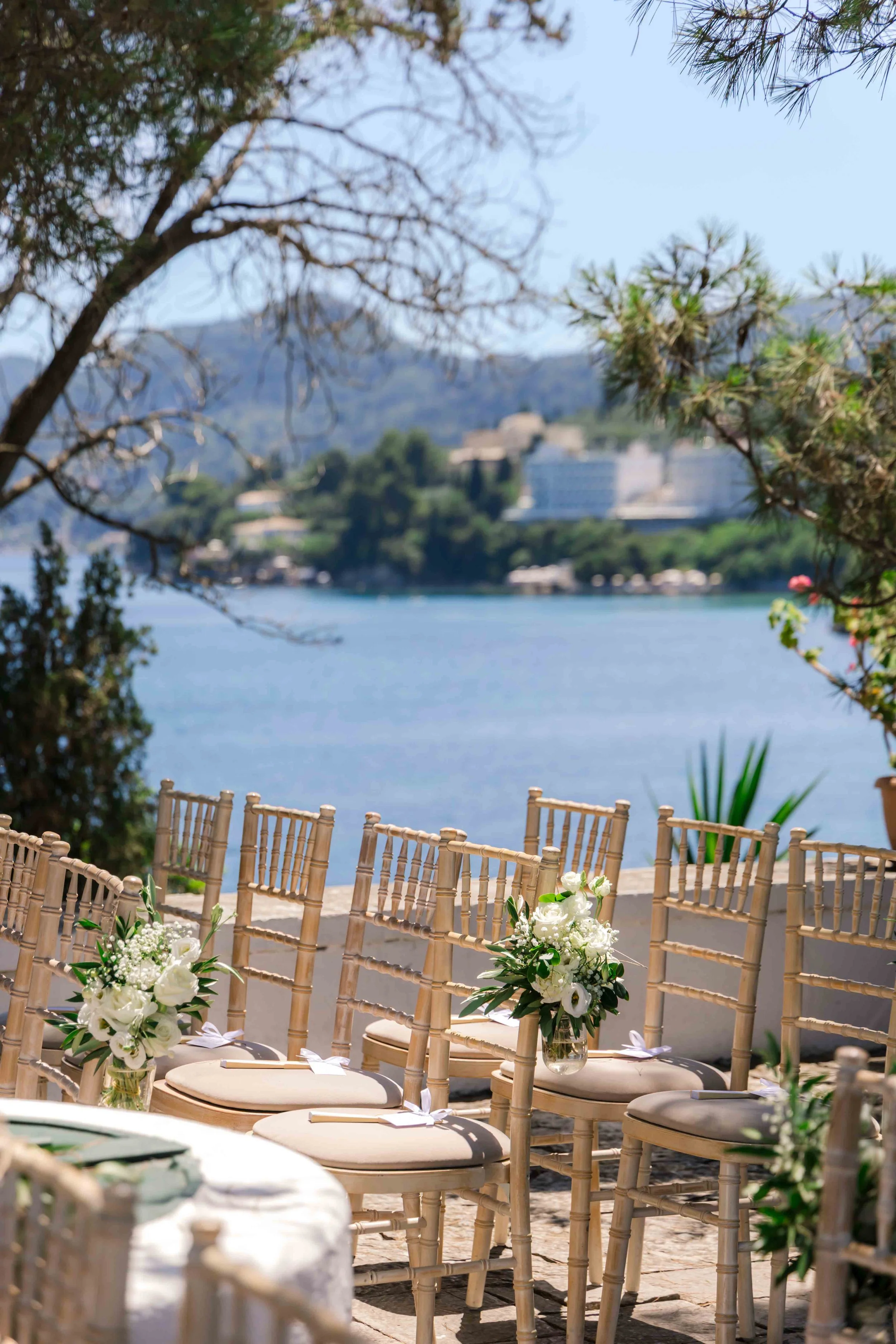 Outdoor wedding ceremony setup with chairs decorated with white flowers, overlooking a body of water and a distant hillside with buildings.