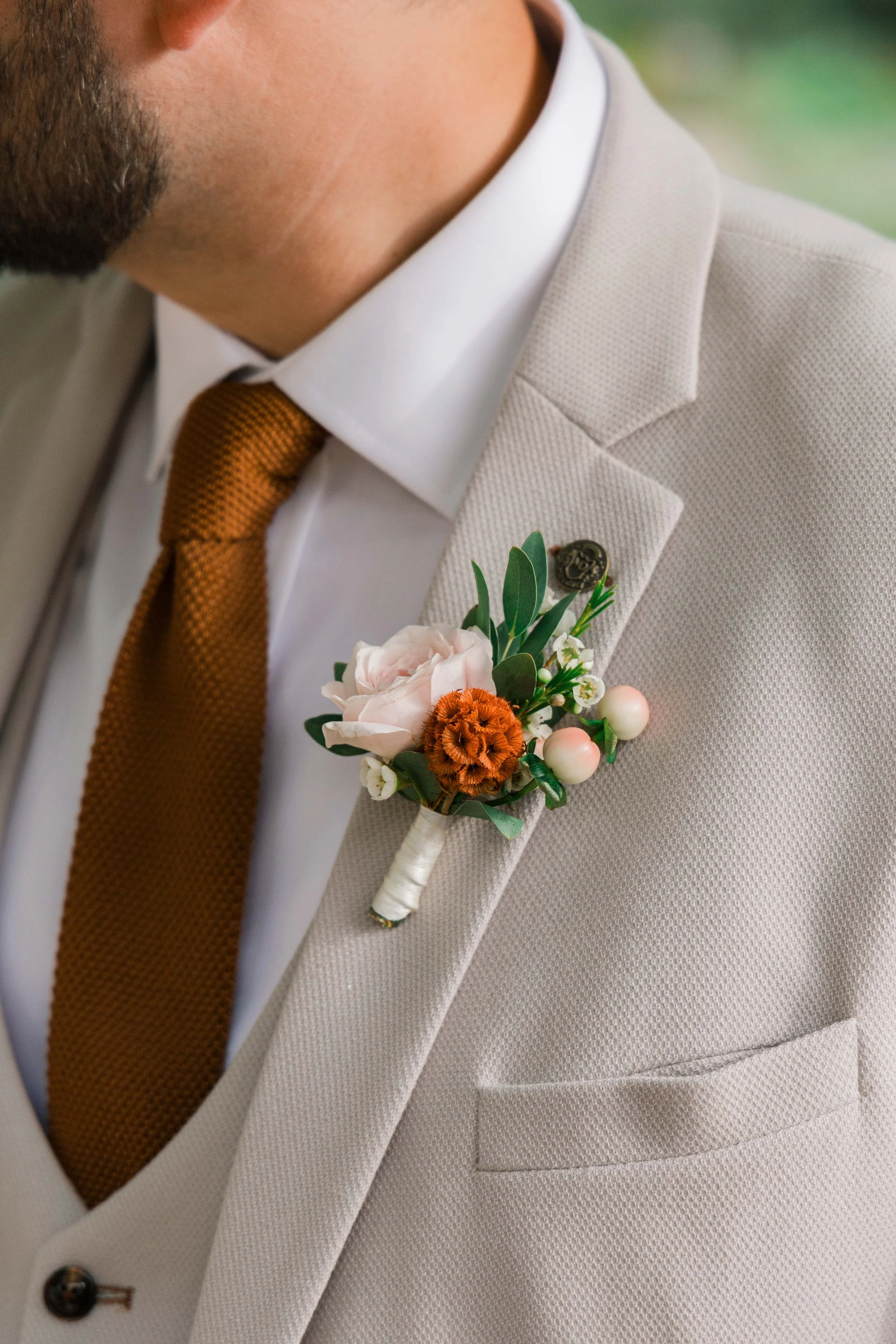 Close-up of a groom's beige suit with a flower boutonniere, a white shirt, and a brown tie, with a small lapel pin.
