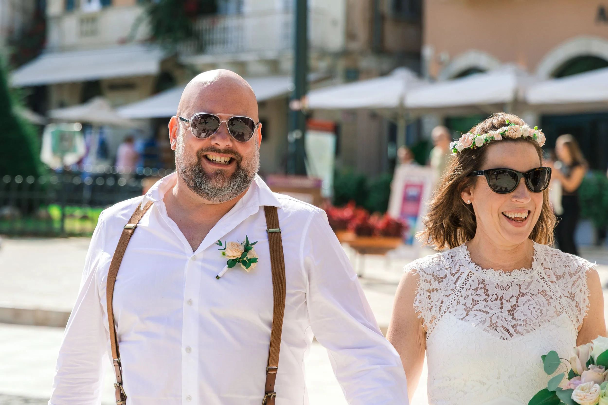 A smiling man and woman dressed in wedding attire, walking outdoors on a sunny day. The man wears sunglasses, a white shirt, suspenders, and a boutonniere. The woman wears sunglasses, a lace dress, and a floral crown. They are holding hands.